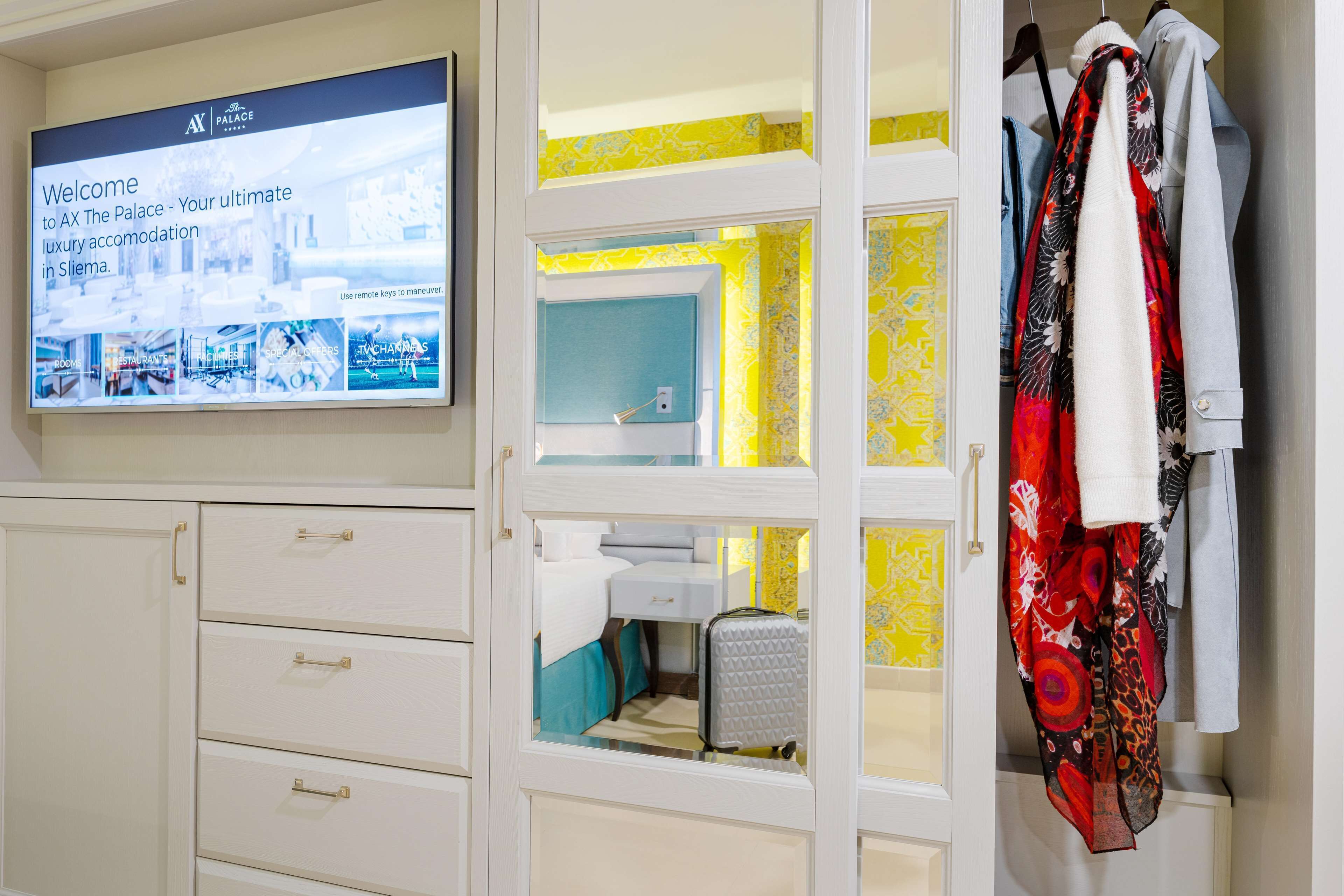 Een hotelkamer met een televisietoestel waarop een welkomstbericht staat, een kledingkast met kleding en een bed met een koffer.