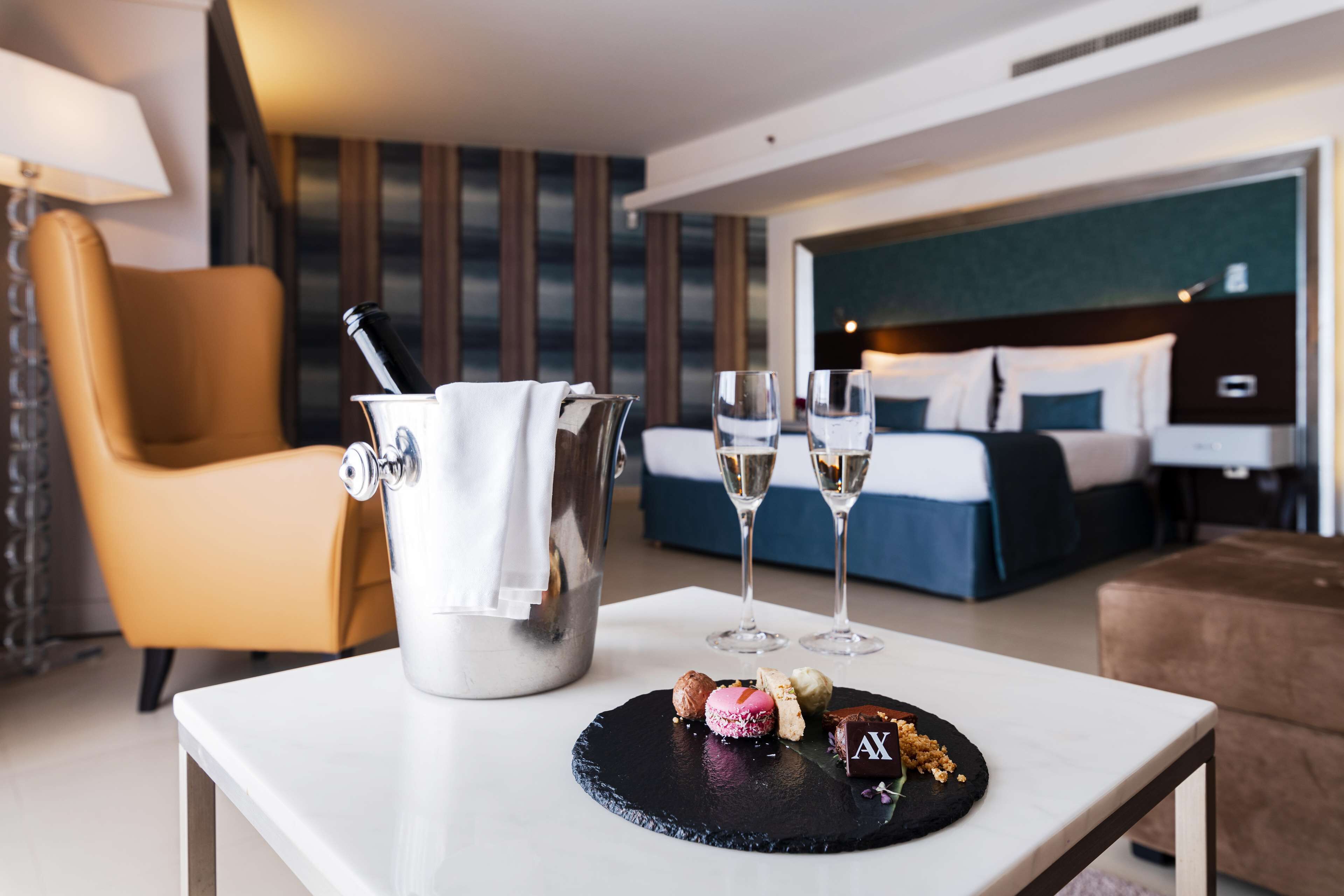 A hotel room with a bed, a bedside table with a pitcher and glasses, and a small table with pastries.
