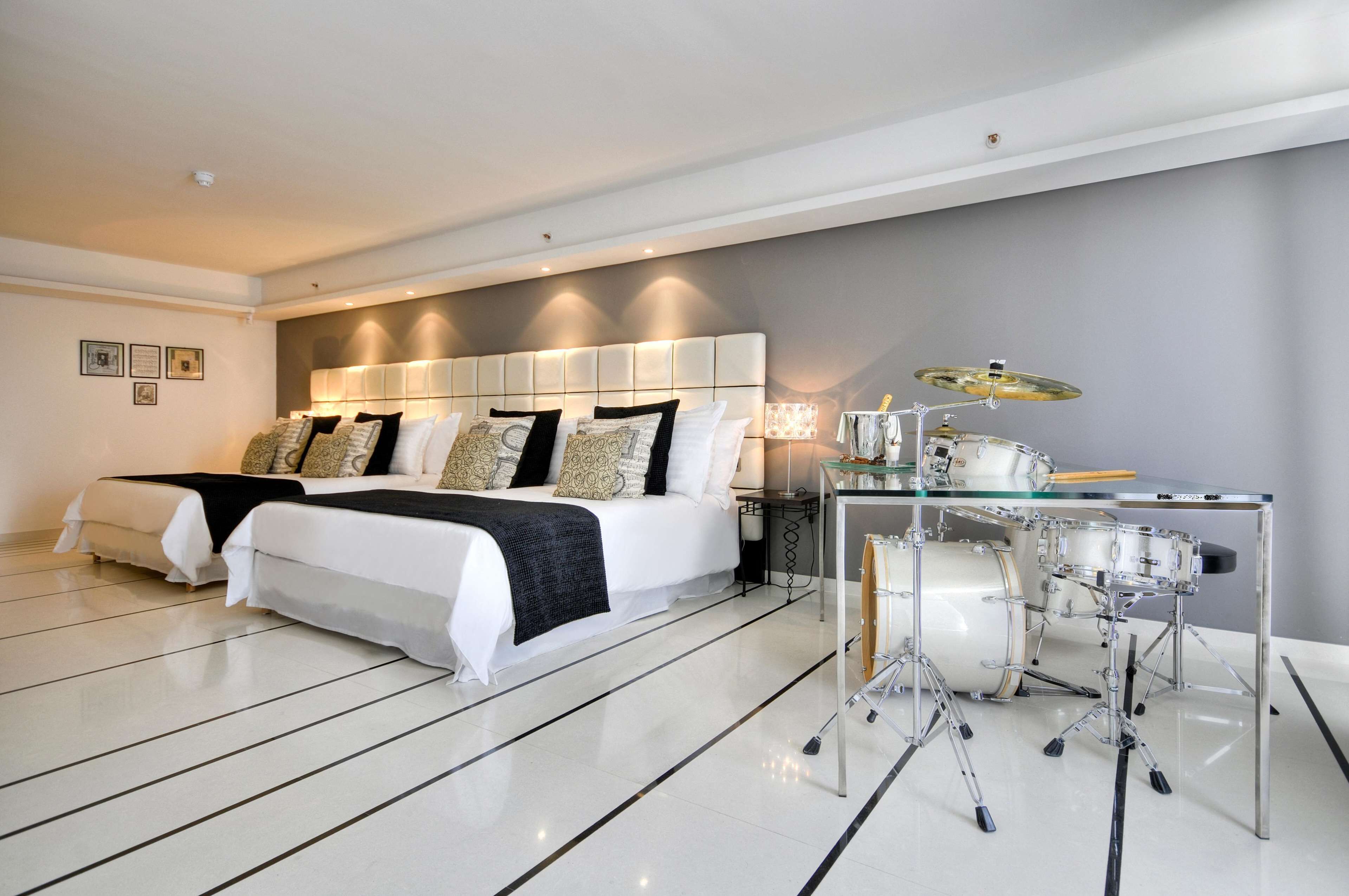 A modern hotel room with a large bed, a separate sofa, a glass table with a drum set, and a well-lit ceiling.