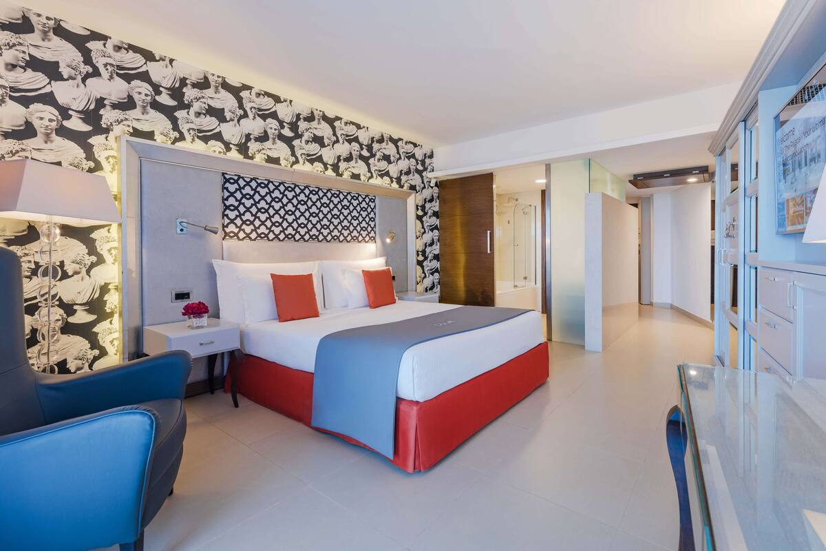 A modern hotel room with a large bed, a bedside table, a chair, and a patterned wall.