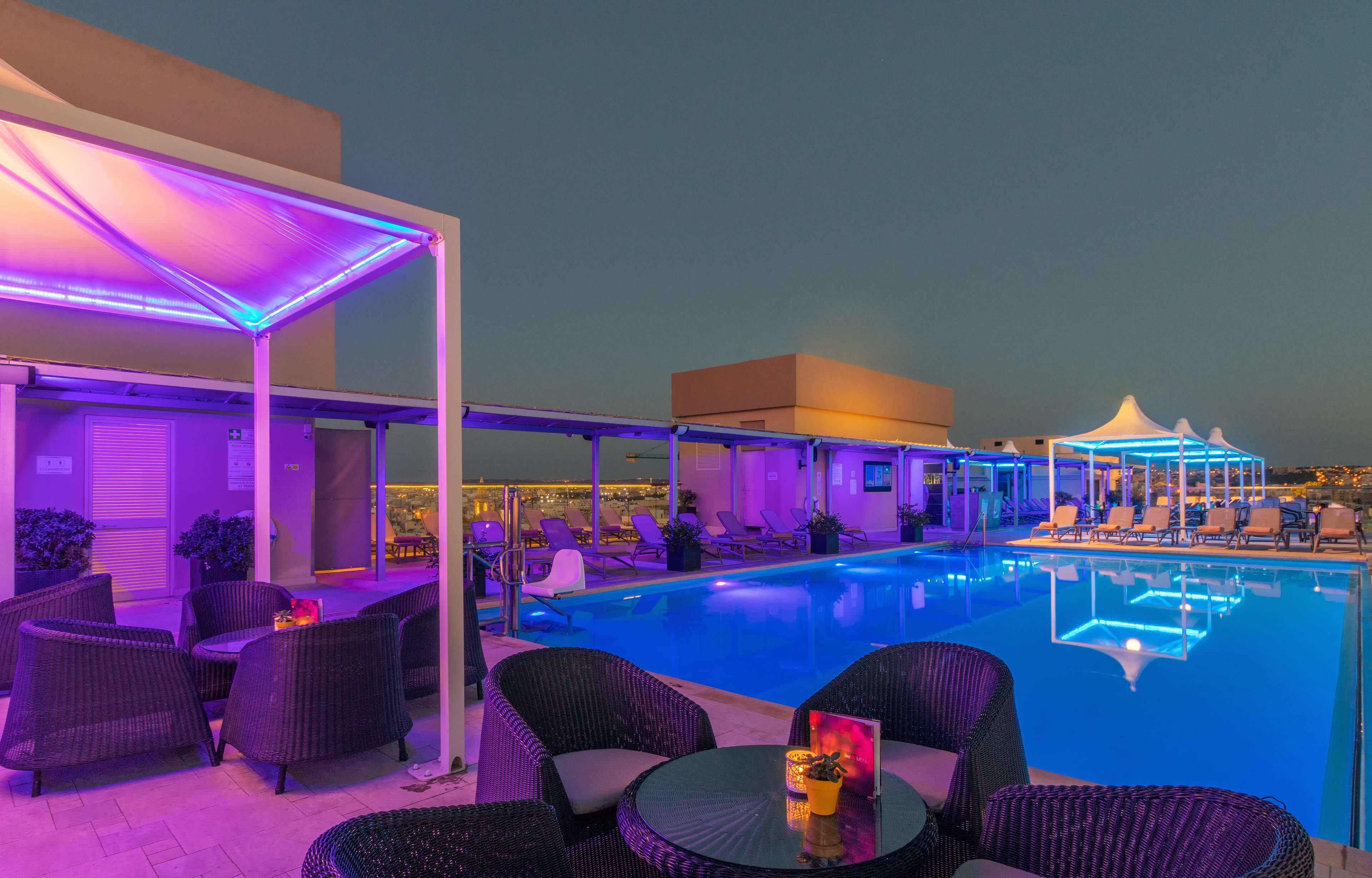 A rooftop pool area with lounge chairs and a table, surrounded by umbrellas and a view of the cityscape.