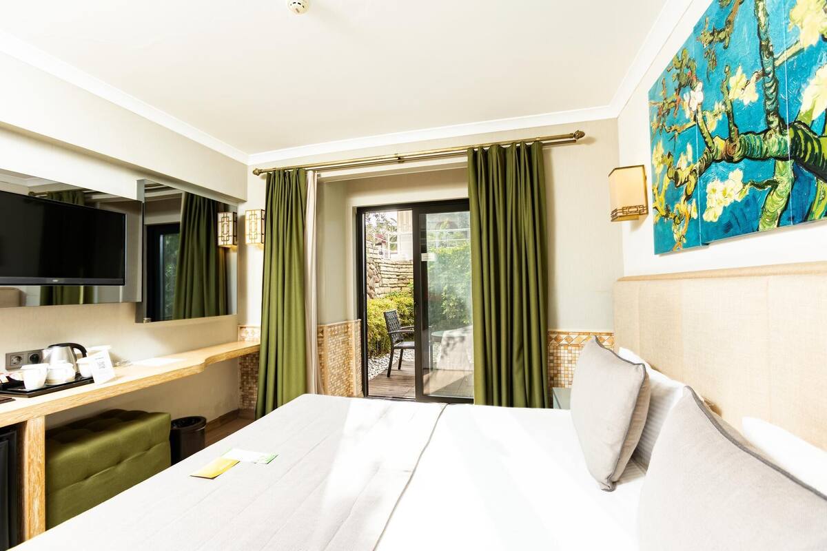 A hotel room with a bed, a desk with a flat-screen TV, a small table with a kettle and cups, and a painting on the wall.
