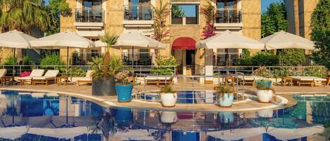 Un espace aménagé au bord de la piscine, avec des chaises longues et des parasols, une piscine qui reflète le bâtiment, et un bâtiment doté de nombreux balcons et d’auvents rouges.