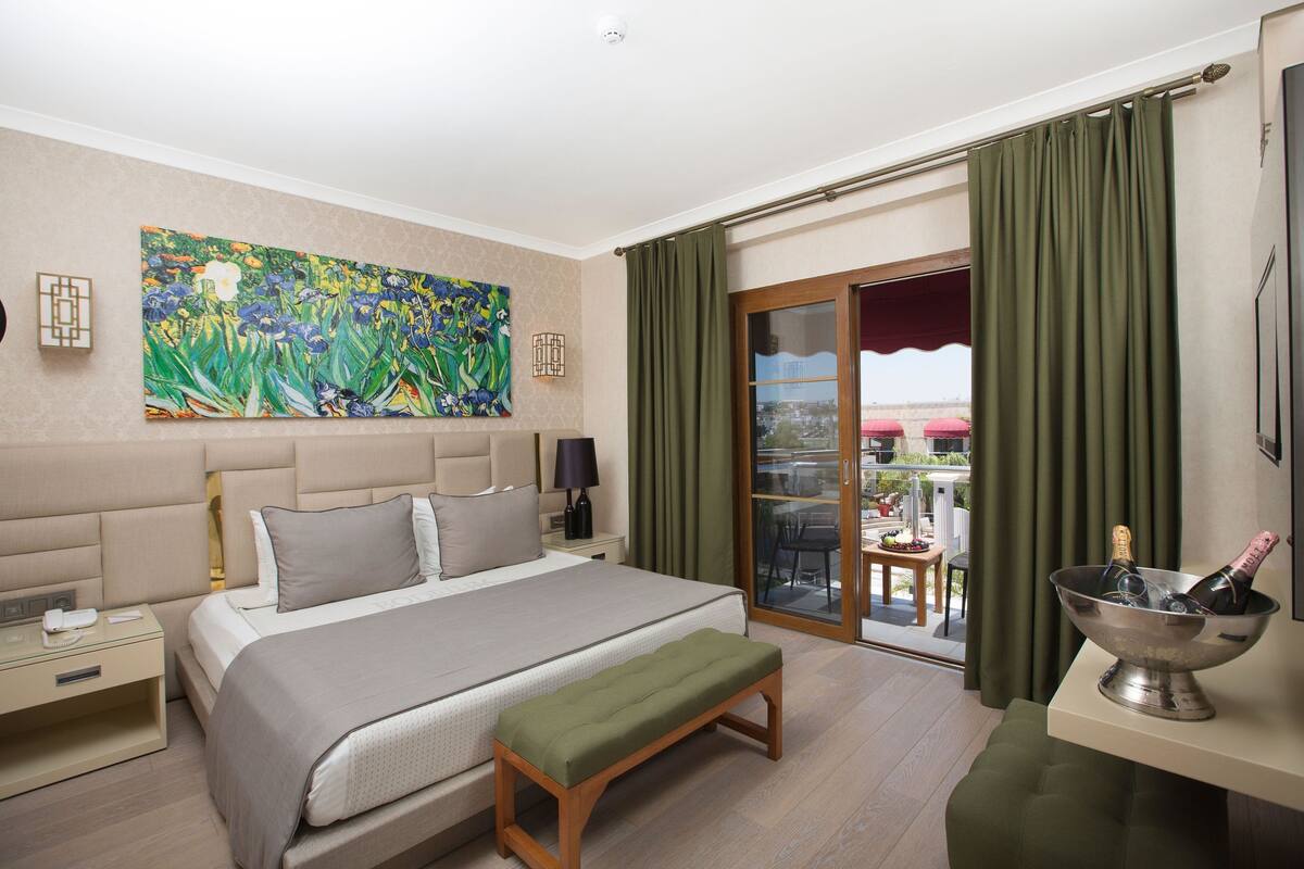 A hotel room with a large bed, a bench, a painting on the wall, a nightstand, a table with a bottle of champagne, and a view of the outside through a sliding door.