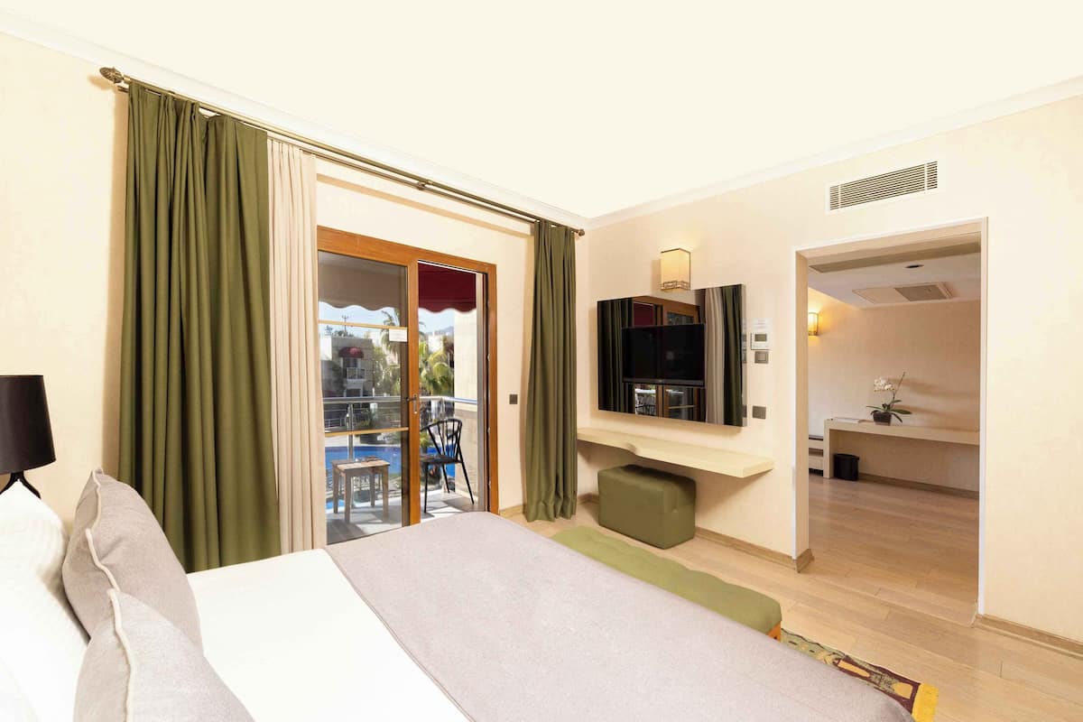 A hotel room with a bed, a TV, a small seating area, and a balcony with curtains.