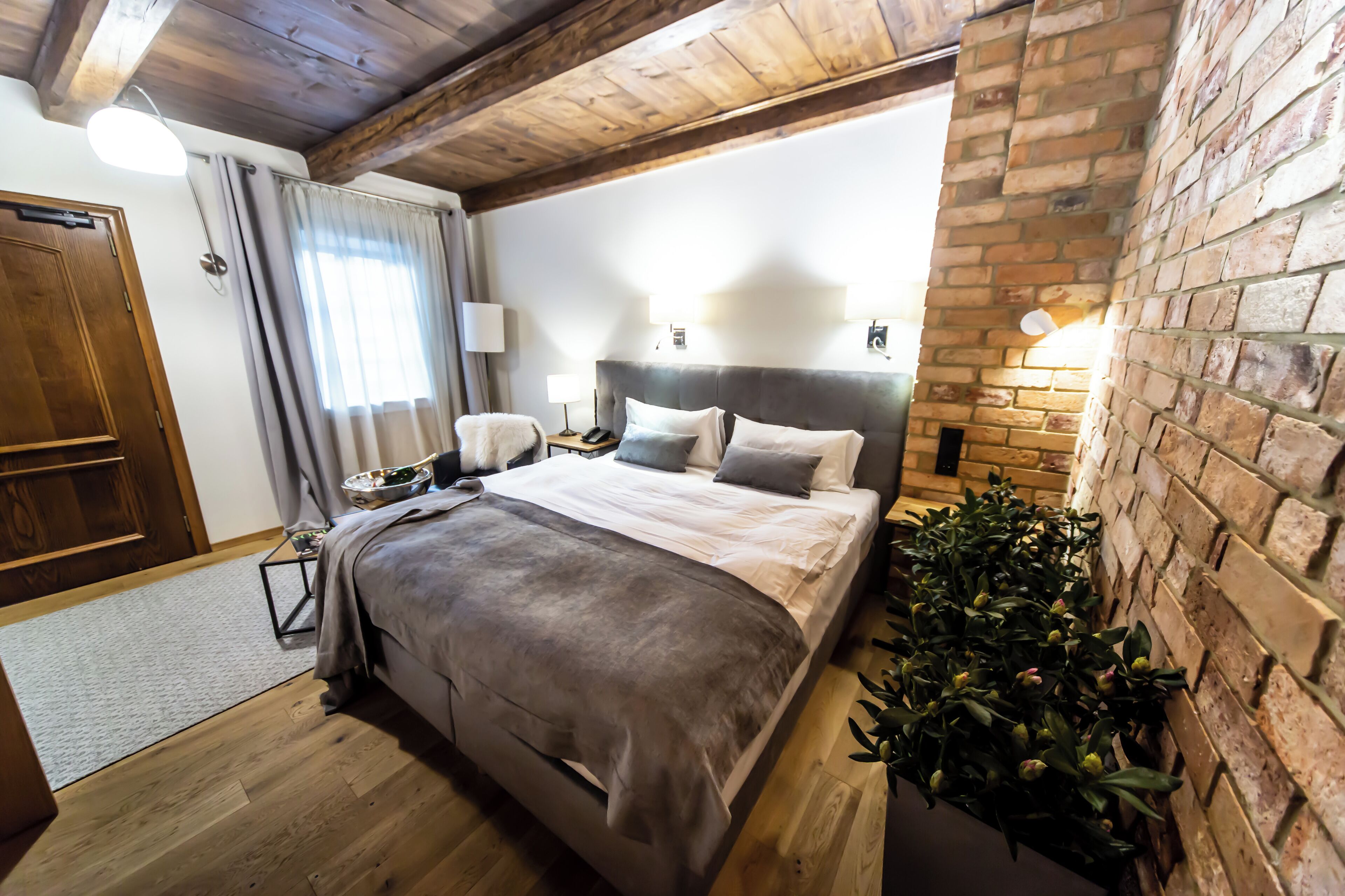 Un dormitorio con techo de madera, pared de ladrillo, cama con manta gris, lámparas de noche y una planta en maceta.