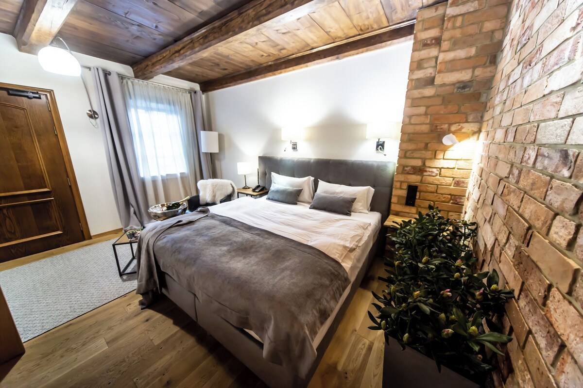 A bedroom with a wooden ceiling, a brick wall, a bed with a grey blanket, bedside lamps, and a potted plant.