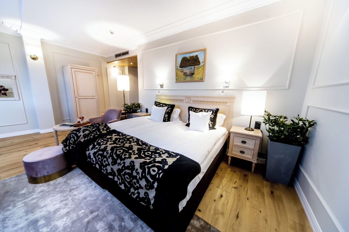 A hotel room with a large bed, bedside tables, a chair, a desk, and a painting on the wall.