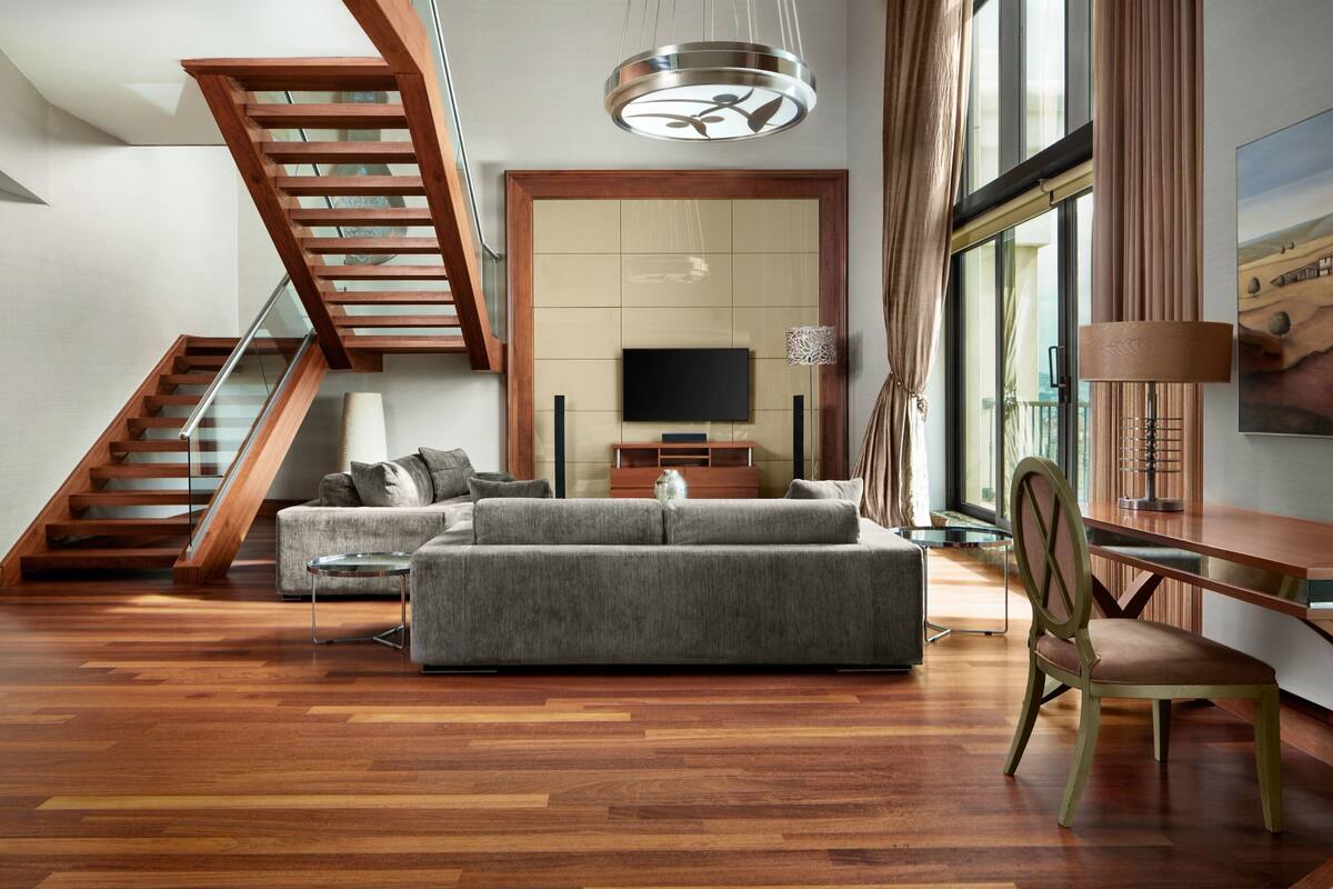 A modern living room with a staircase, a large sofa, a dining table, and a TV mounted on the wall.