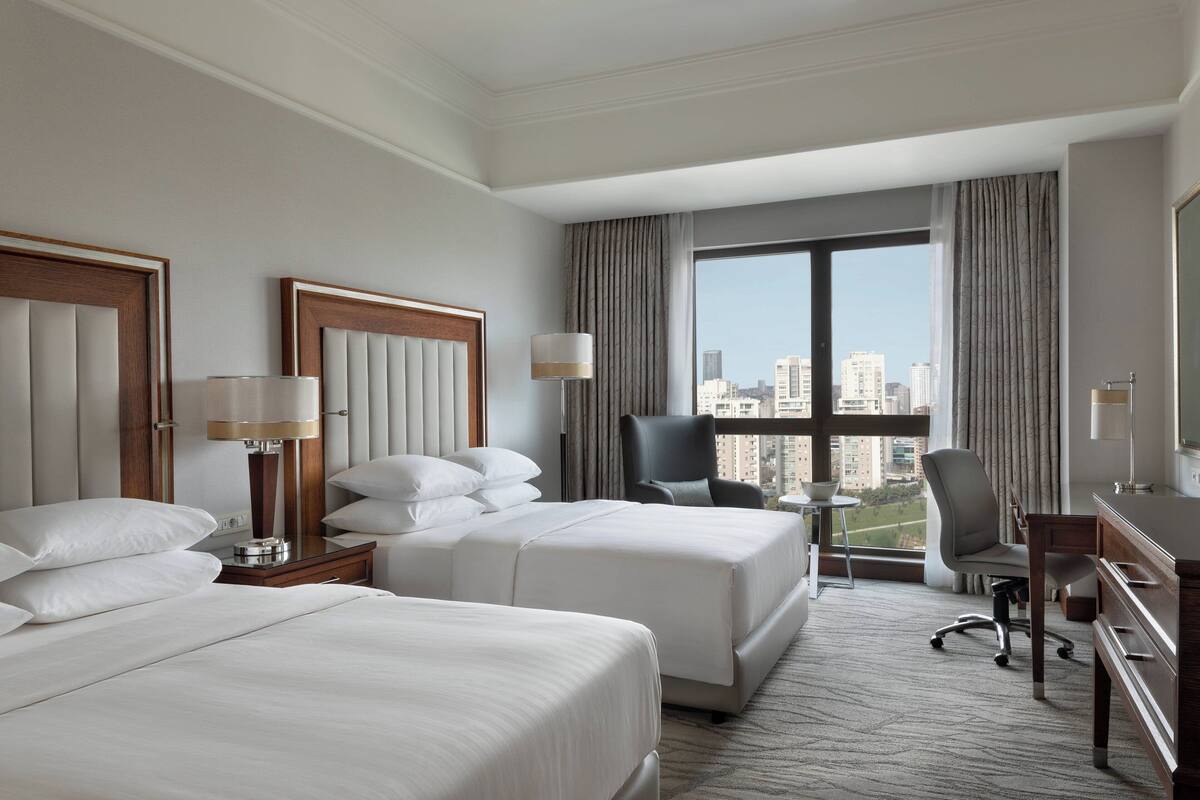 A hotel room with two beds, a desk, a chair, and a large window offering a city view.