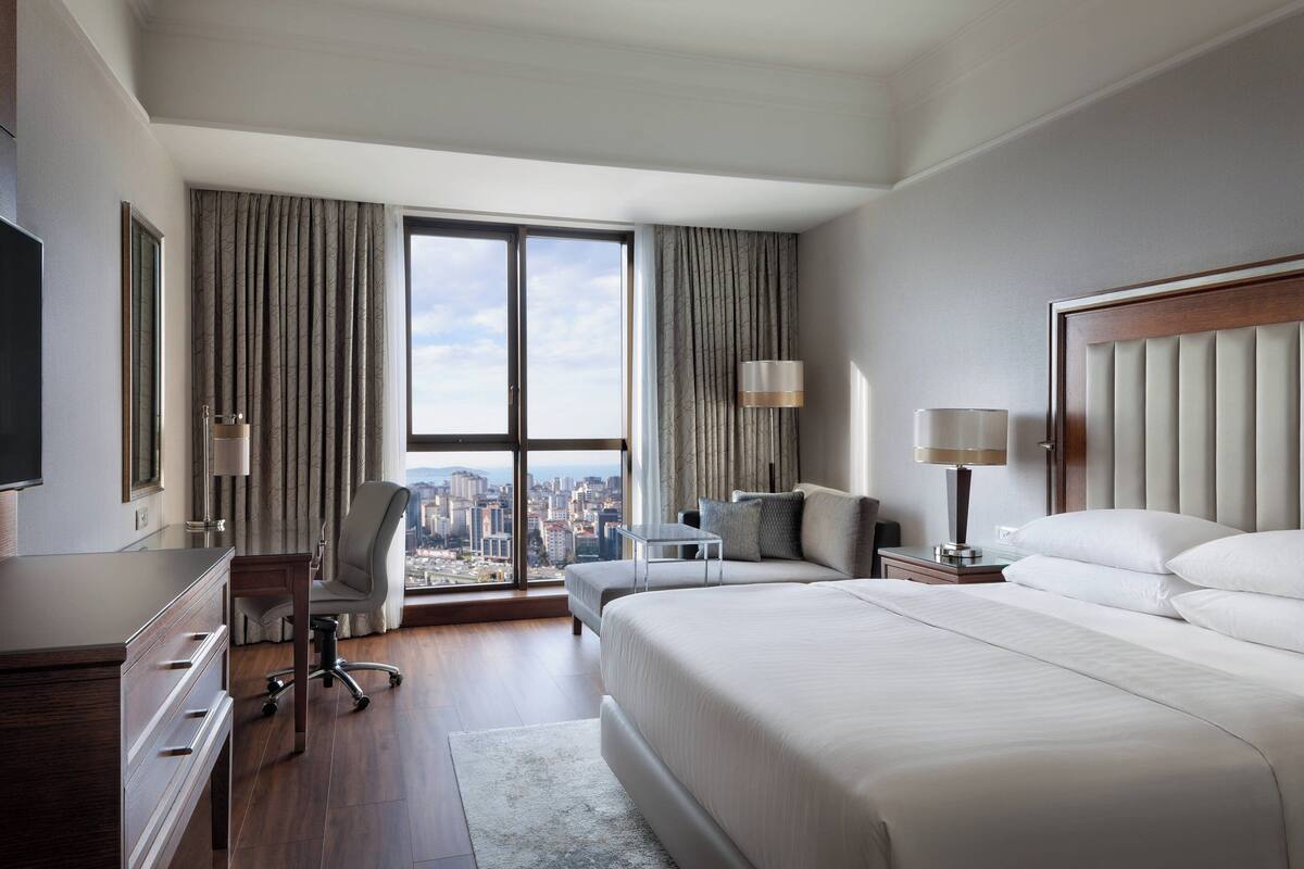 A hotel room with a large bed, a desk, a chair, and a view of the city through the window.