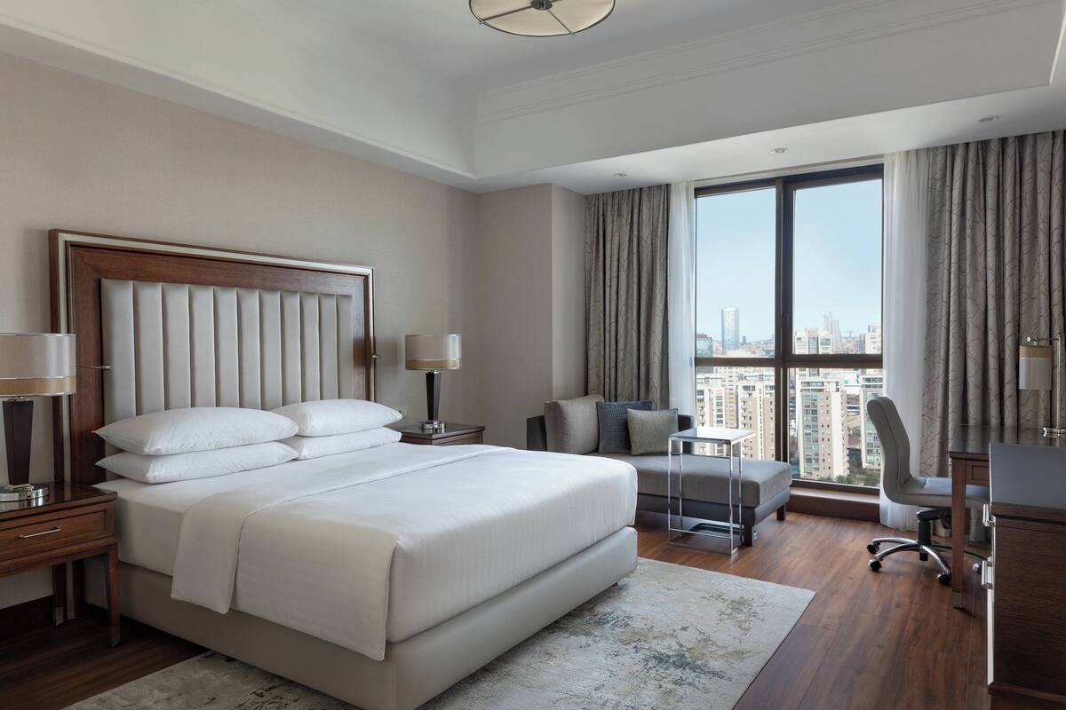 A modern hotel room with a large bed, a sofa, a desk, and a view of the city.