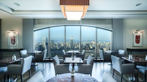 A modern dining area with large windows offering a city view. Tables and chairs are arranged for dining.