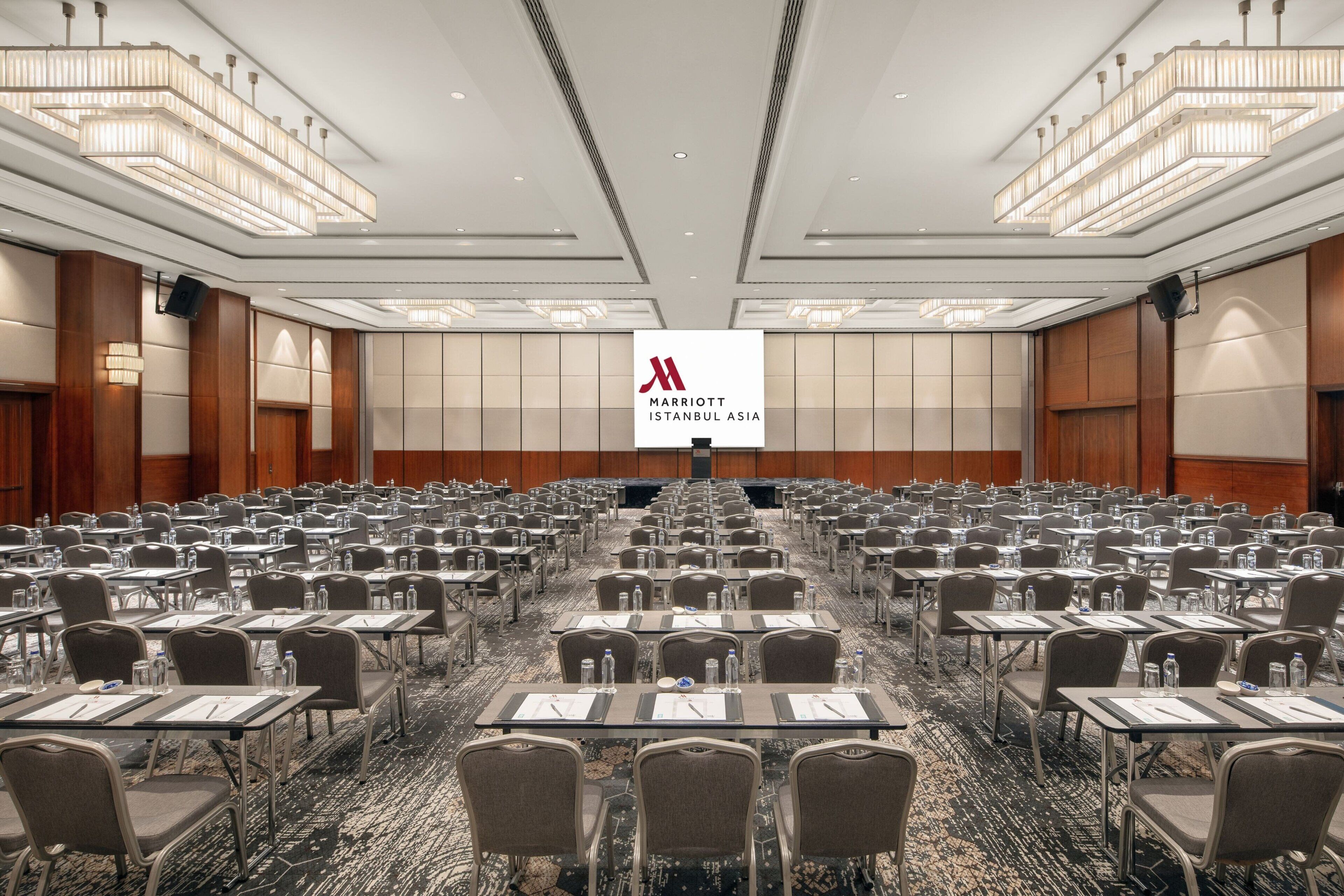 Ein großer Konferenzraum mit Tischen und Stühlen in Reihen, einem zentralen Bildschirm, der das Logo des Marriott Istanbul Asia zeigt, und einer modernen Kristallleuchter.