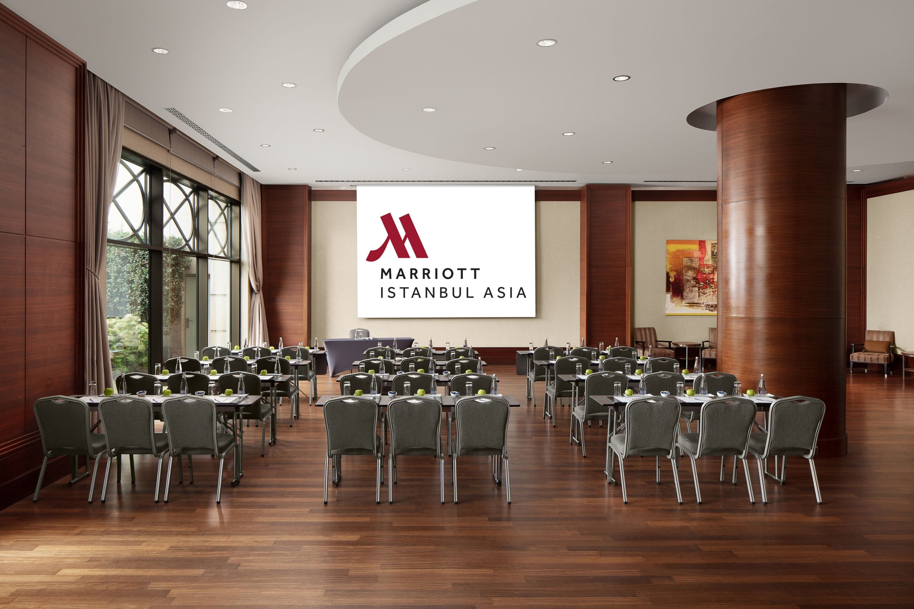 Une salle de conférence à l’hôtel Marriott Istanbul Asia, équipée de plusieurs tables et chaises, de grandes fenêtres et ornée d’un logo bien visible sur le mur.
