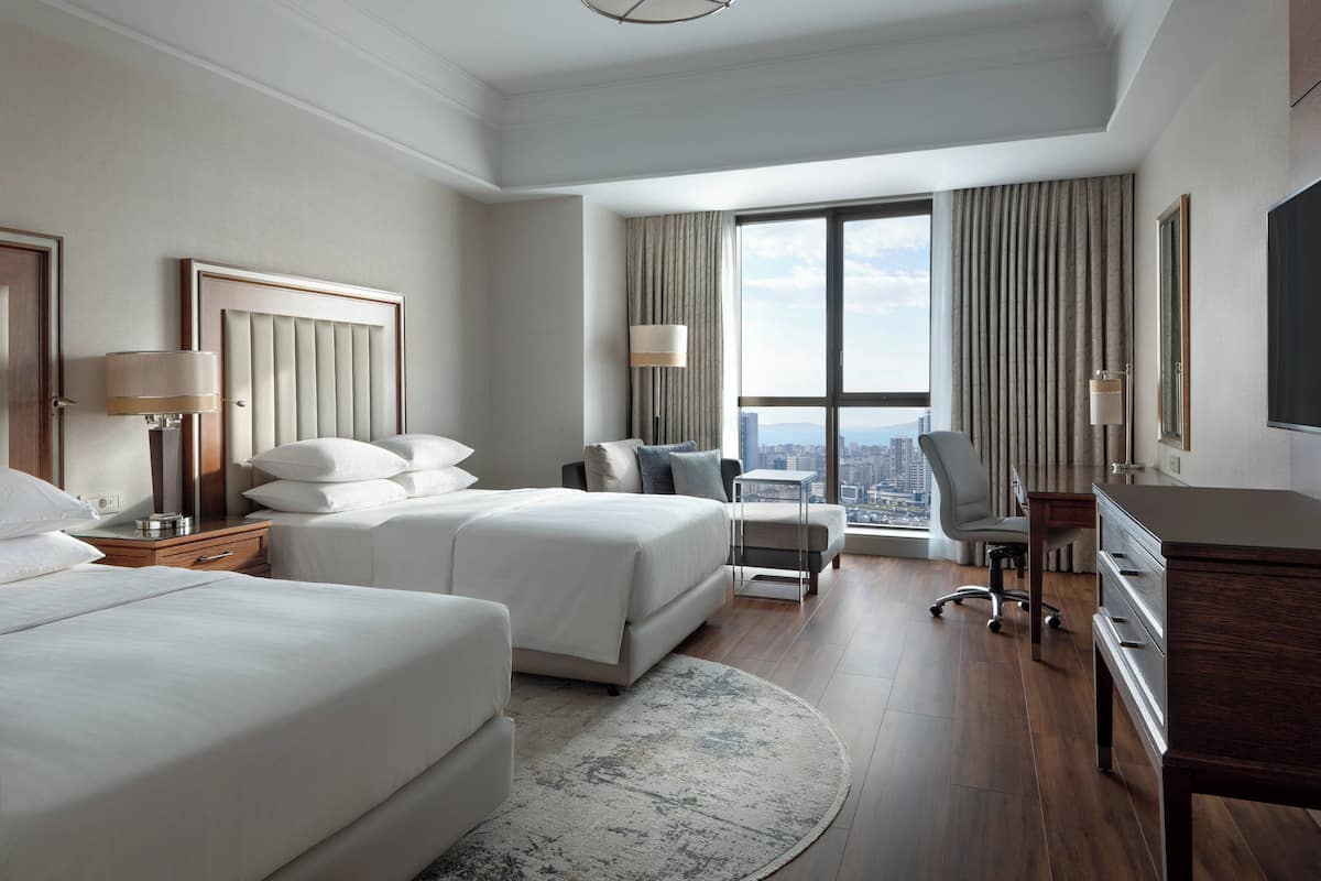 A hotel room with two beds, a desk, a chair, and a large window with a city view.