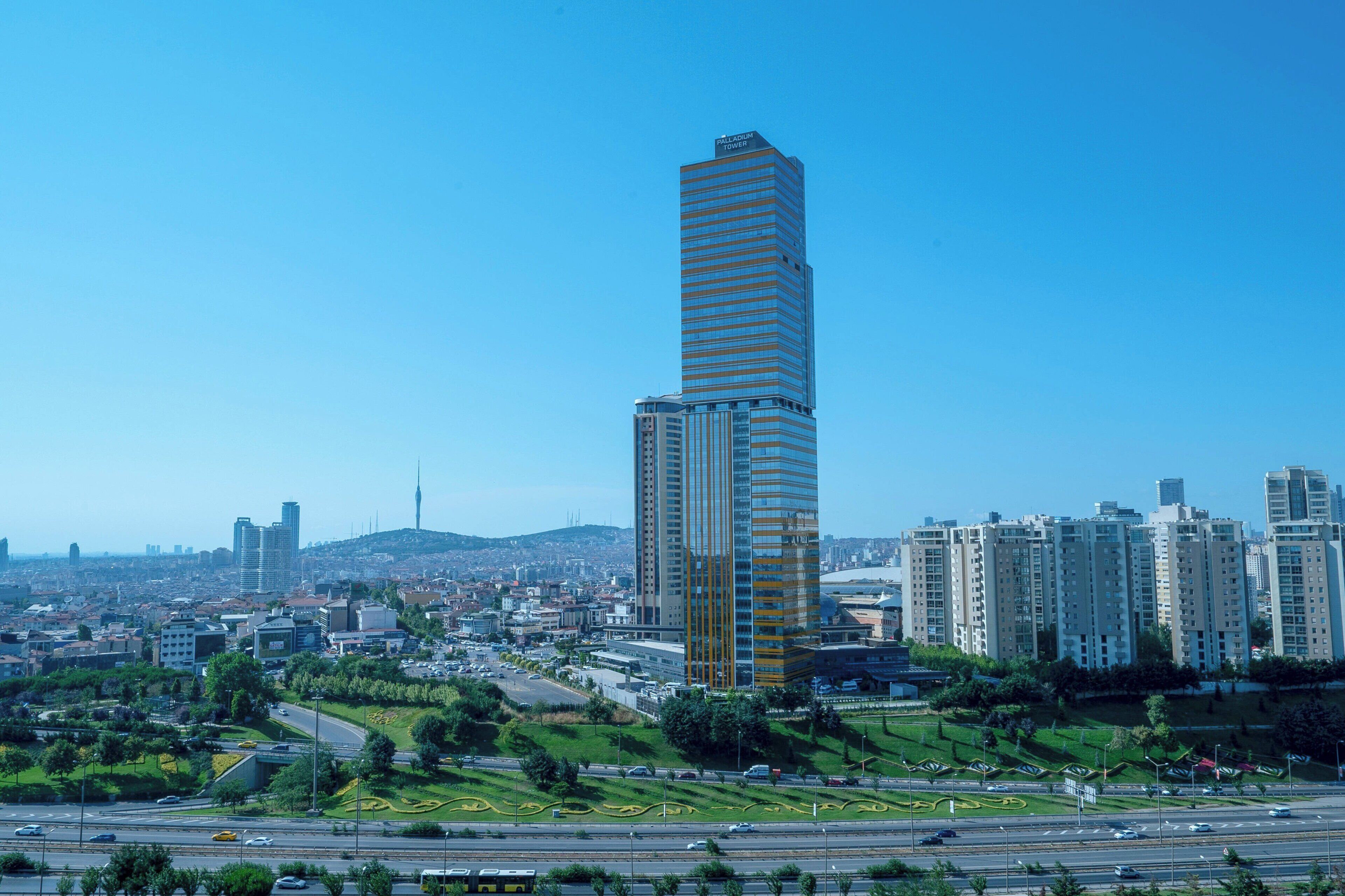 Un gratte-ciel moderne et imposant, doté d’une façade à rayures originales, s’élève au milieu d’un paysage urbain composé de différents bâtiments, sous un ciel bleu dégagé.