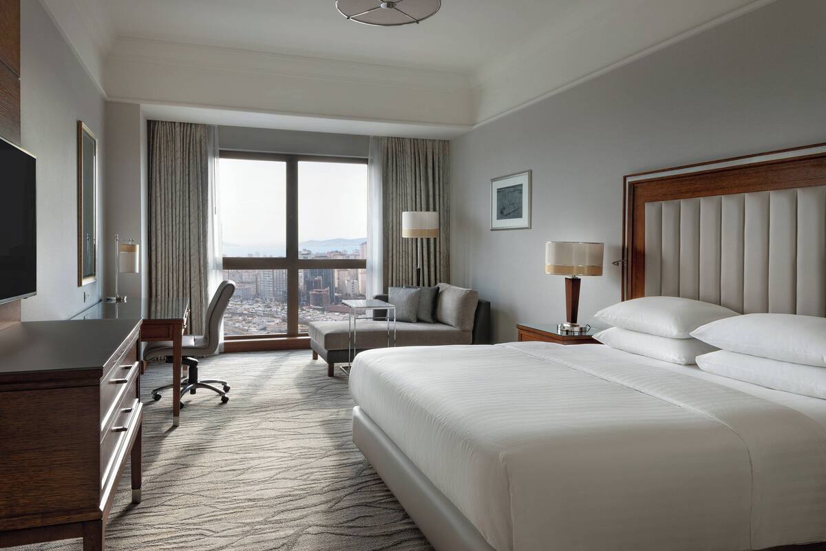 A hotel room with a large bed, a desk, a chair, a small table, and a view of the city.