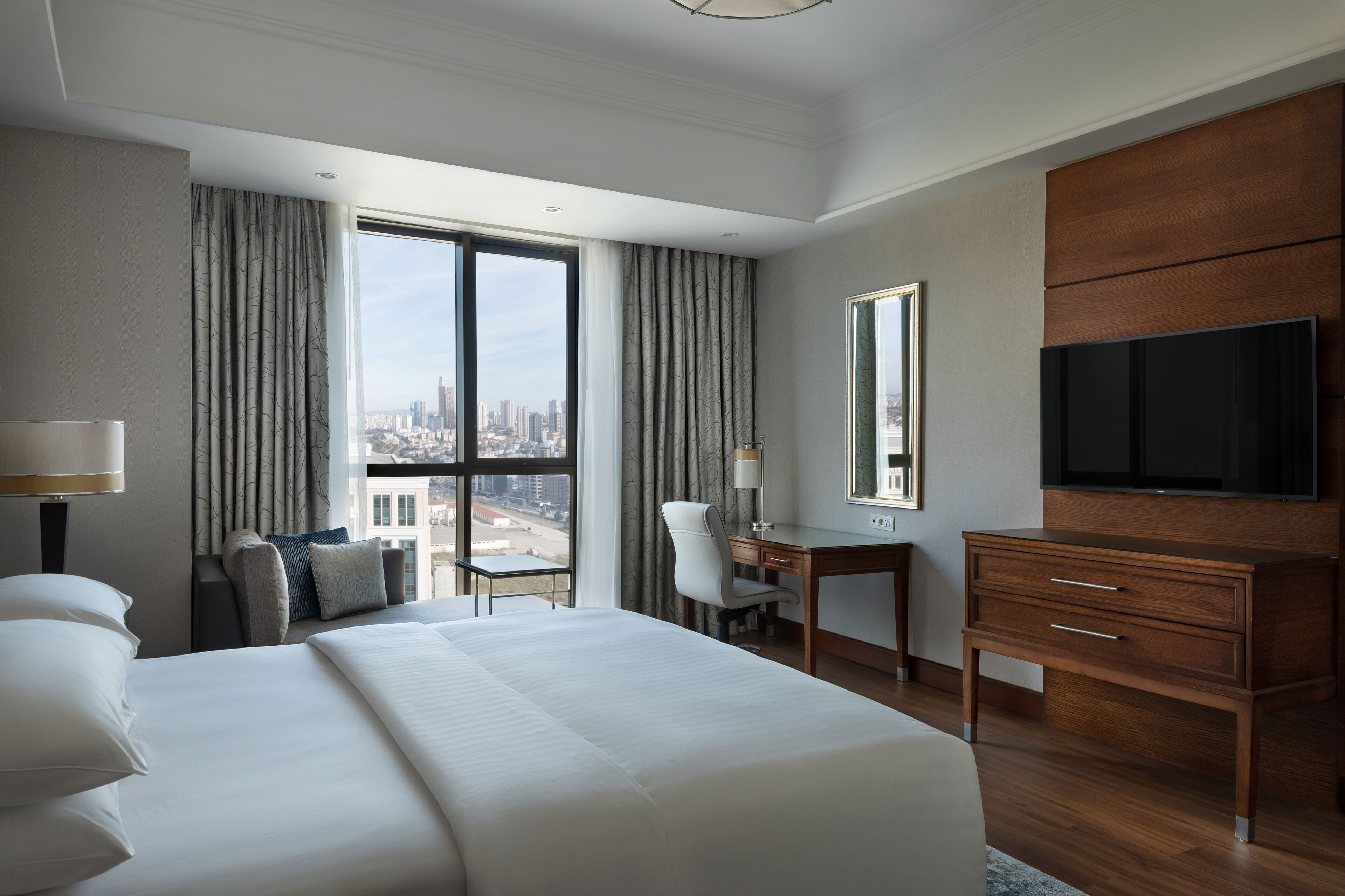 Ein Hotelzimmer mit einem großen Bett, einem Schreibtisch, einem Stuhl, einem Fernseher und Blick auf die Stadt.