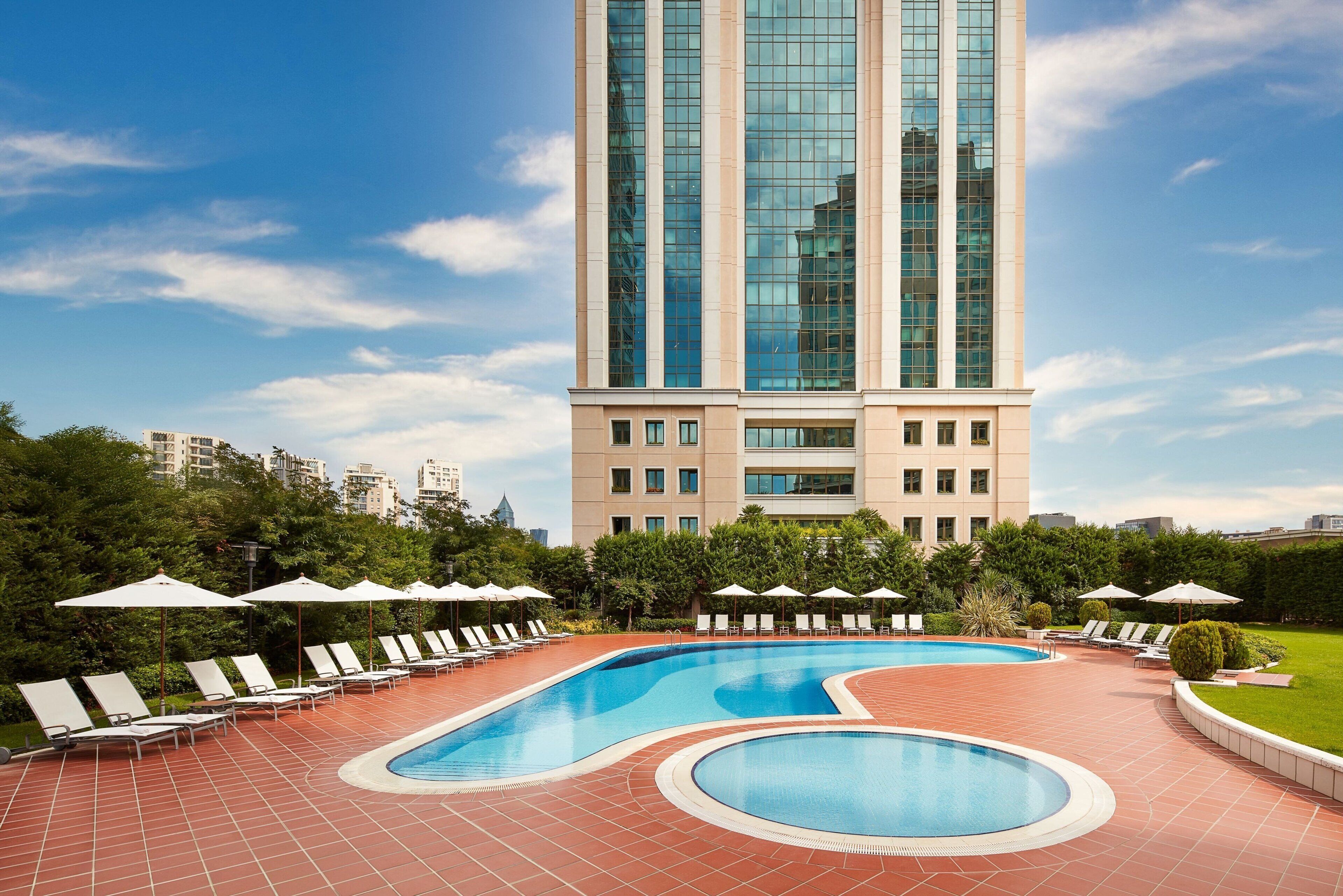 A swimming pool with lounge chairs and umbrellas, surrounded by a brick patio and greenery, with a tall building in the background.