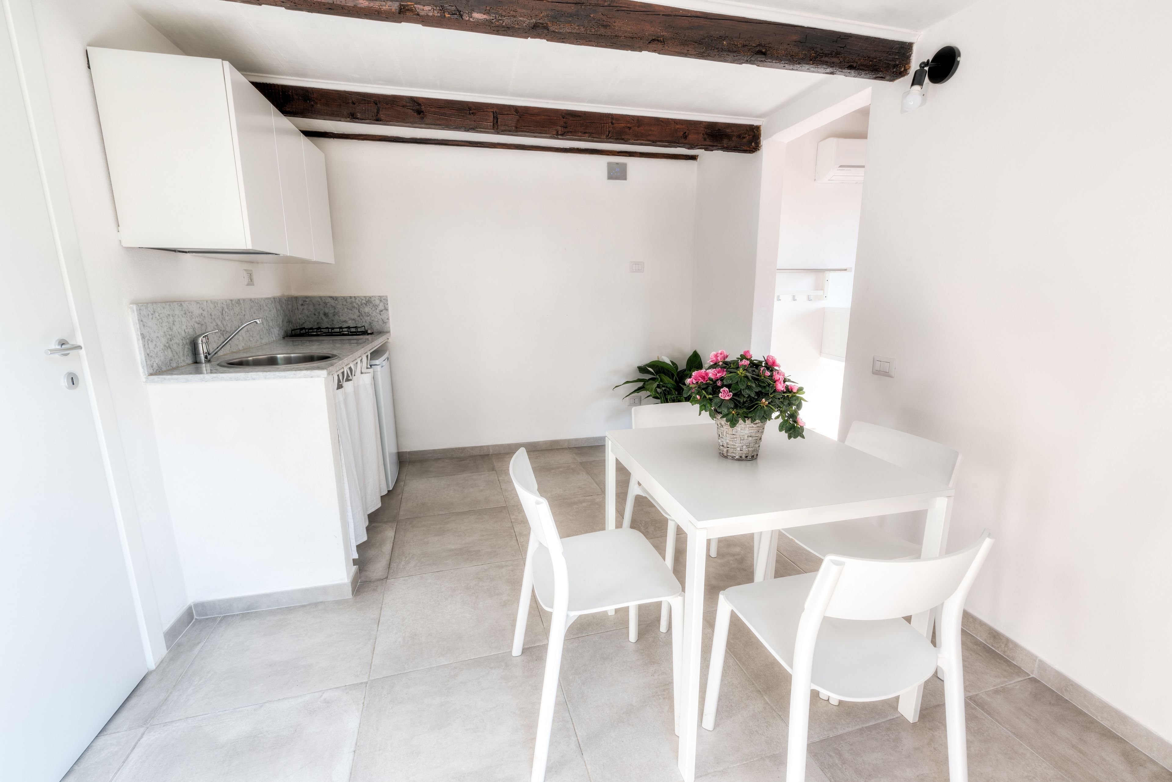 Una cocina moderna con armarios blancos, encimera de granito y pileta integrada. Hay un comedor con una mesa y sillas blancas, y una planta en maceta con flores rosas.