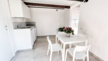 Una cocina moderna con armarios blancos, encimera de granito y pileta integrada. Hay un comedor con una mesa y sillas blancas, y una planta en maceta con flores rosas.