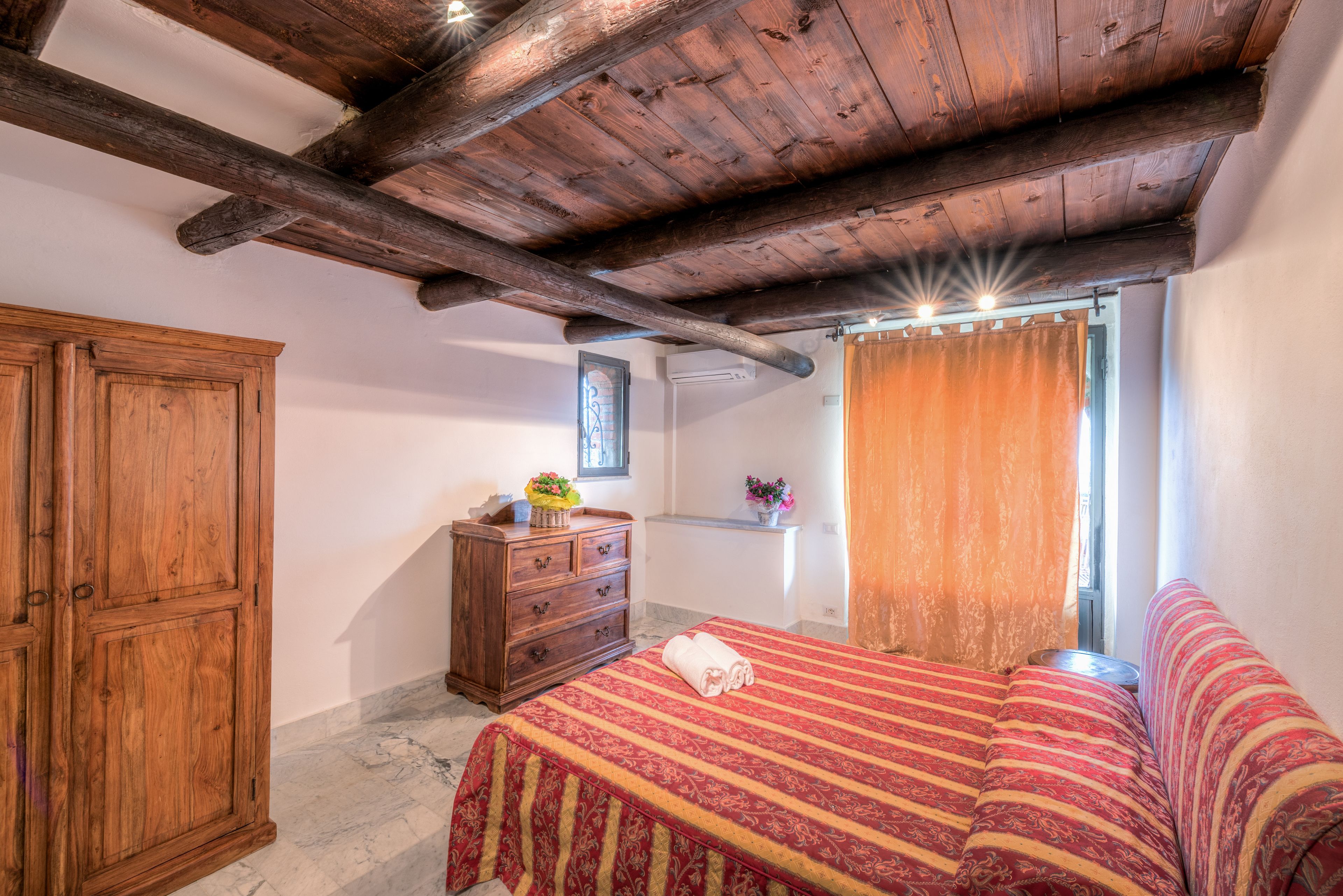 A bedroom with a wooden ceiling, a bed with a red and yellow striped bedspread, a wooden dresser, and a window with curtains.