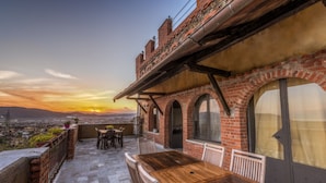 A rooftop terrace with a wooden table and chairs, brick walls, and large windows overlooking a cityscape at sunset. - Ippotur Medieval Resort (Castelnuovo Magra)