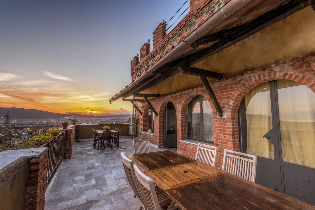 A rooftop terrace with a wooden table and chairs, brick walls, and large windows overlooking a cityscape at sunset.