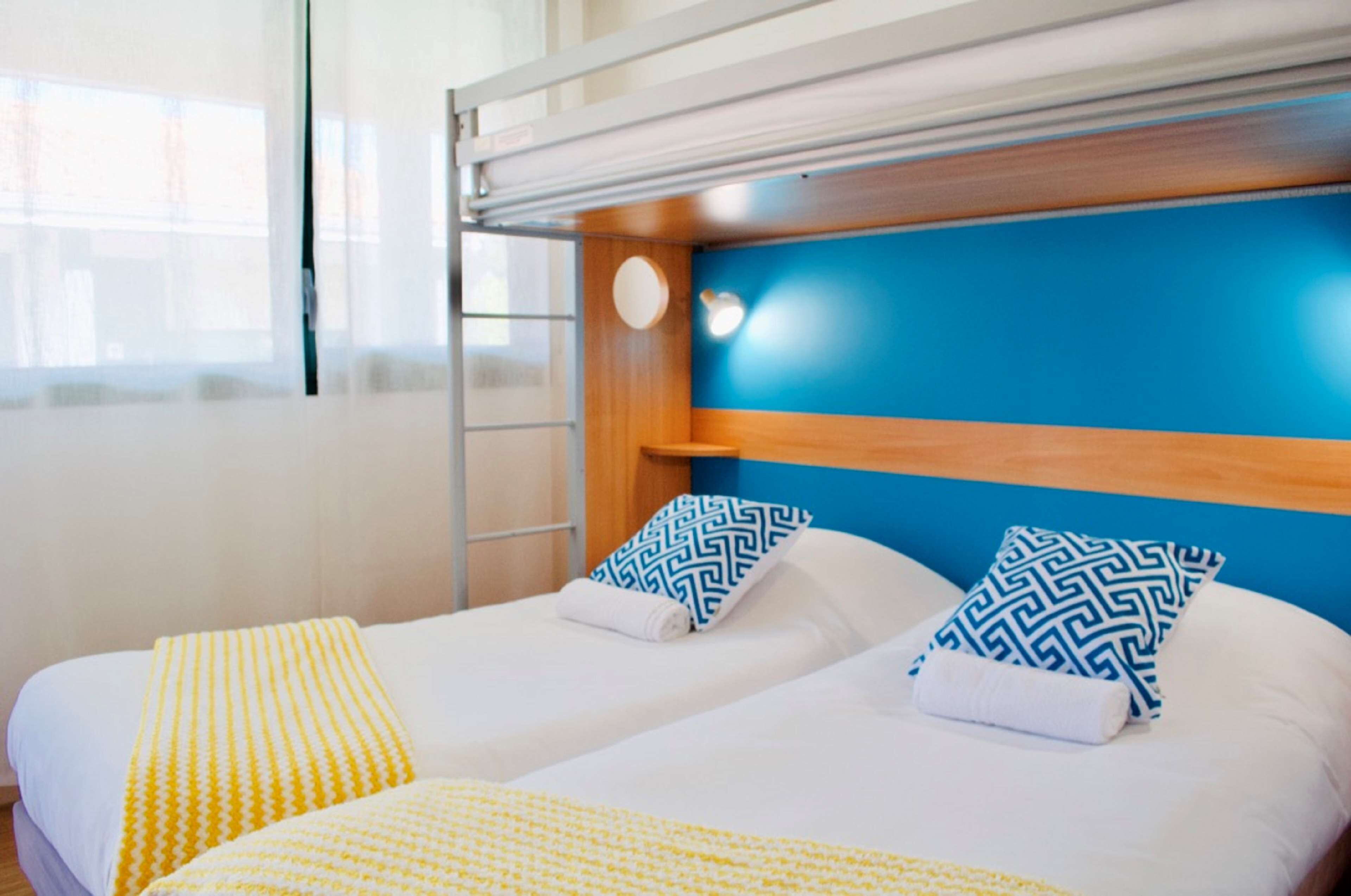 A double room with two beds, each with a white mattress and a yellow blanket. There is a blue headboard with a round light fixture.