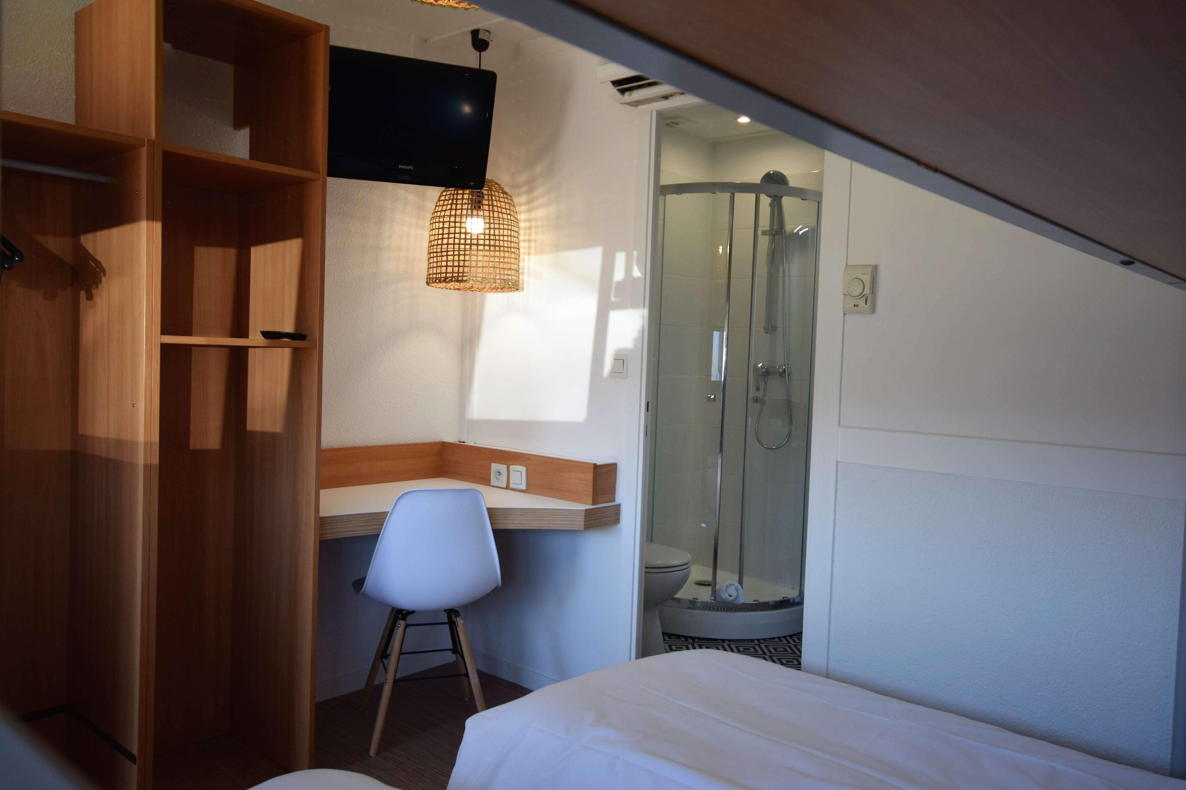 Habitación de hotel con cama, escritorio con silla, armario, zona de ducha y televisor montado en la pared.