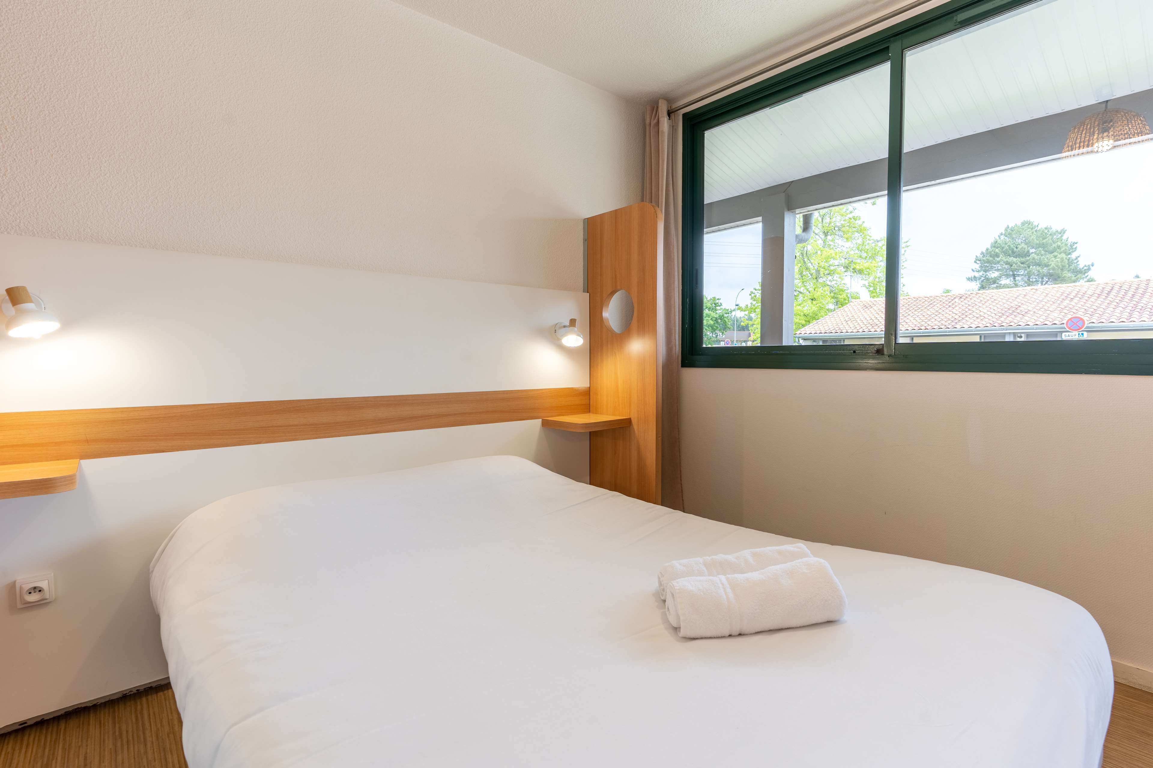 Une chambre d’hôtel avec un lit simple, une tête de lit en bois, une fenêtre donnant sur des immeubles et un luminaire fixé au mur.