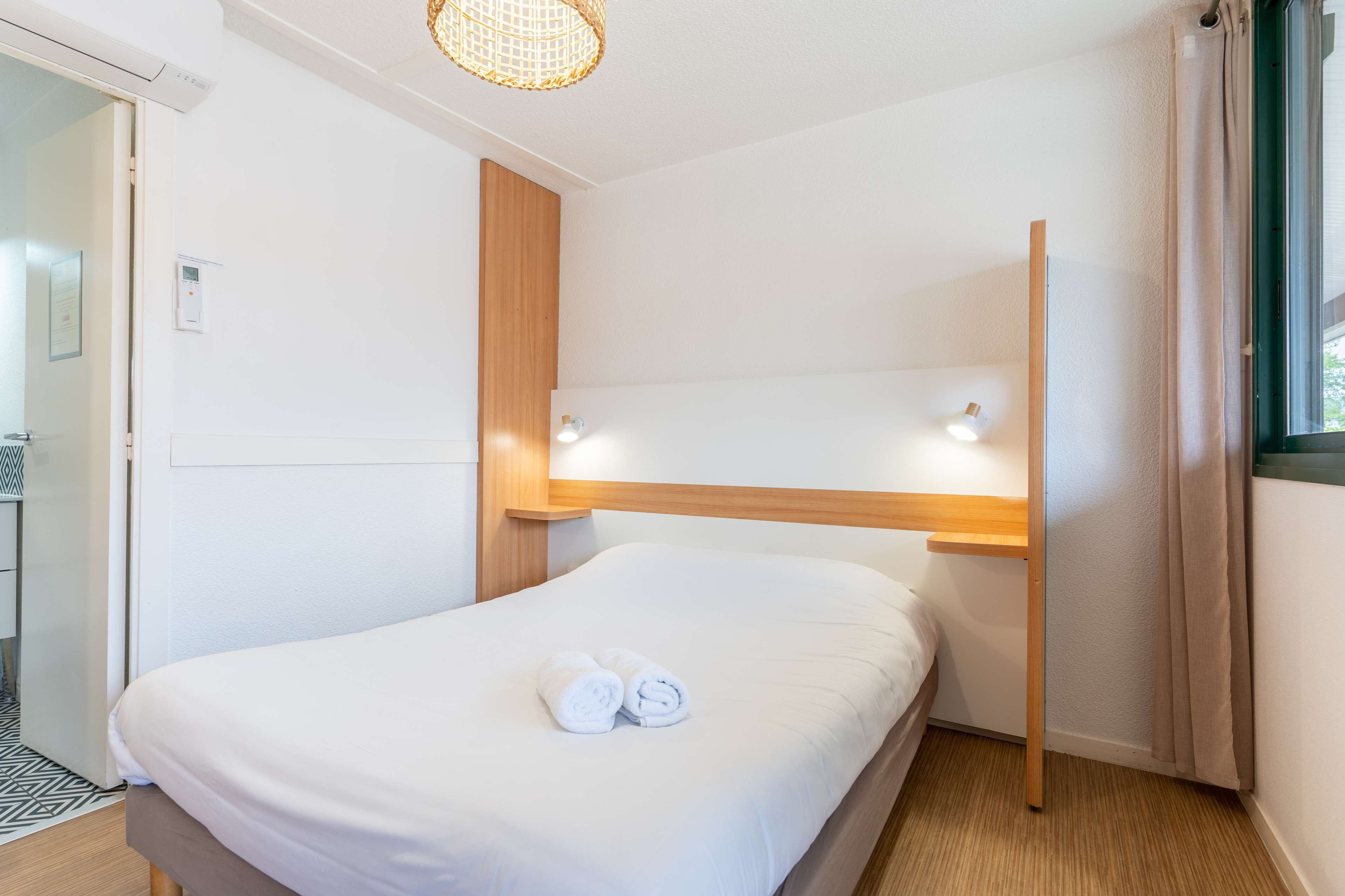 Un lit bien fait, avec du linge de lit blanc et des serviettes pliées, une tête de lit en bois et une table de chevet avec une lampe.