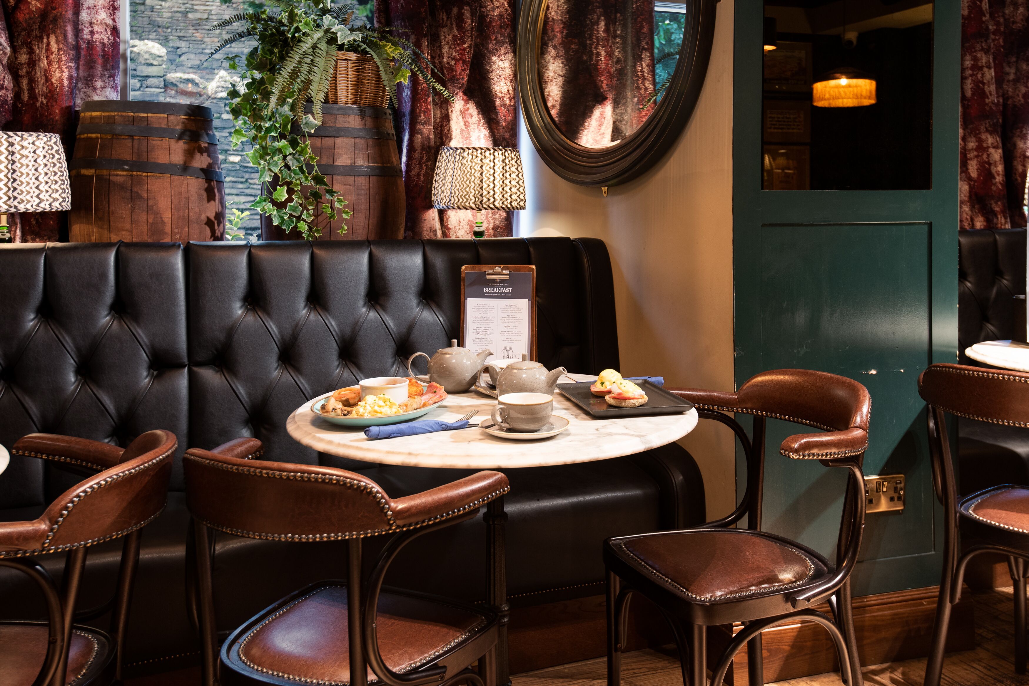 A restaurant with a black leather booth, wooden chairs, a round table with breakfast items, and a menu board.