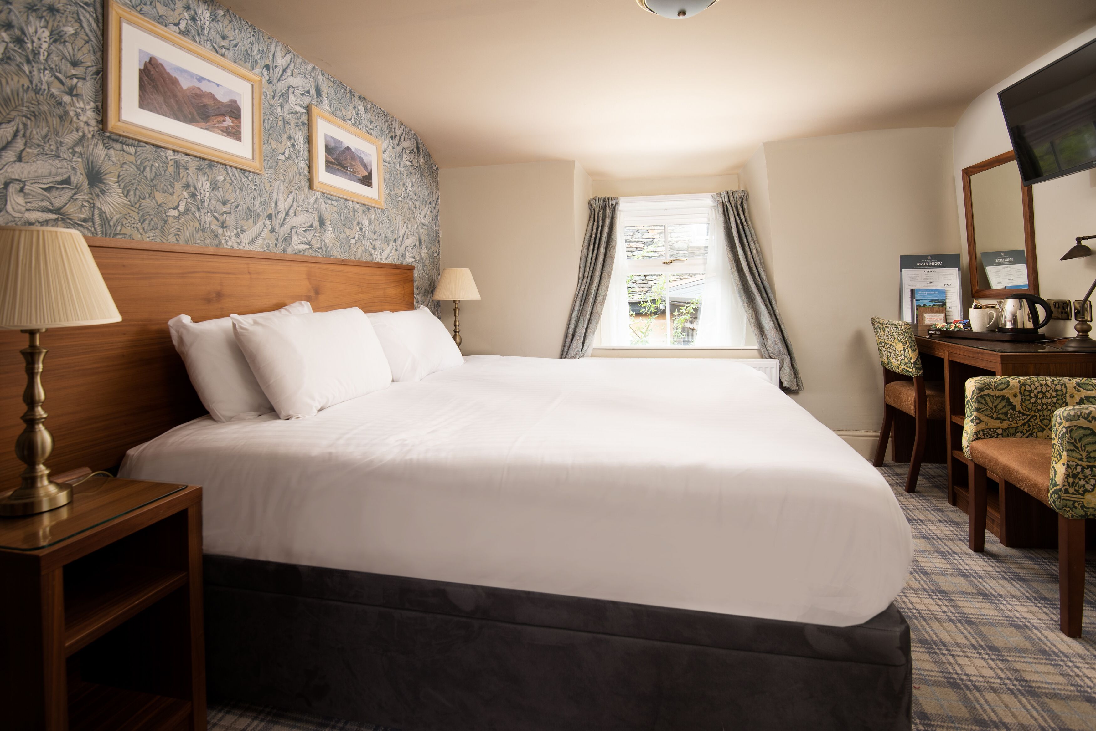 A hotel room with a large bed, a wooden nightstand, a desk with a chair, a television, and framed pictures on the wall.