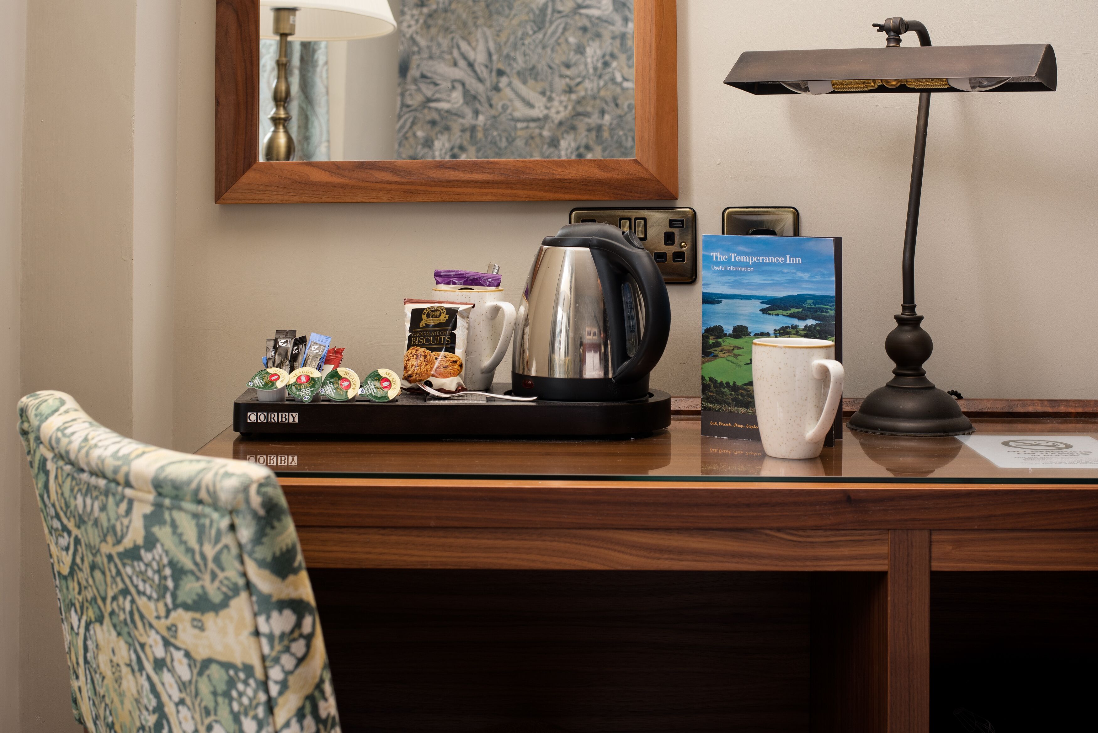 A wooden desk with a kettle, a coffee maker, a book titled 'The Temperance Inn', a framed picture, and a lamp.