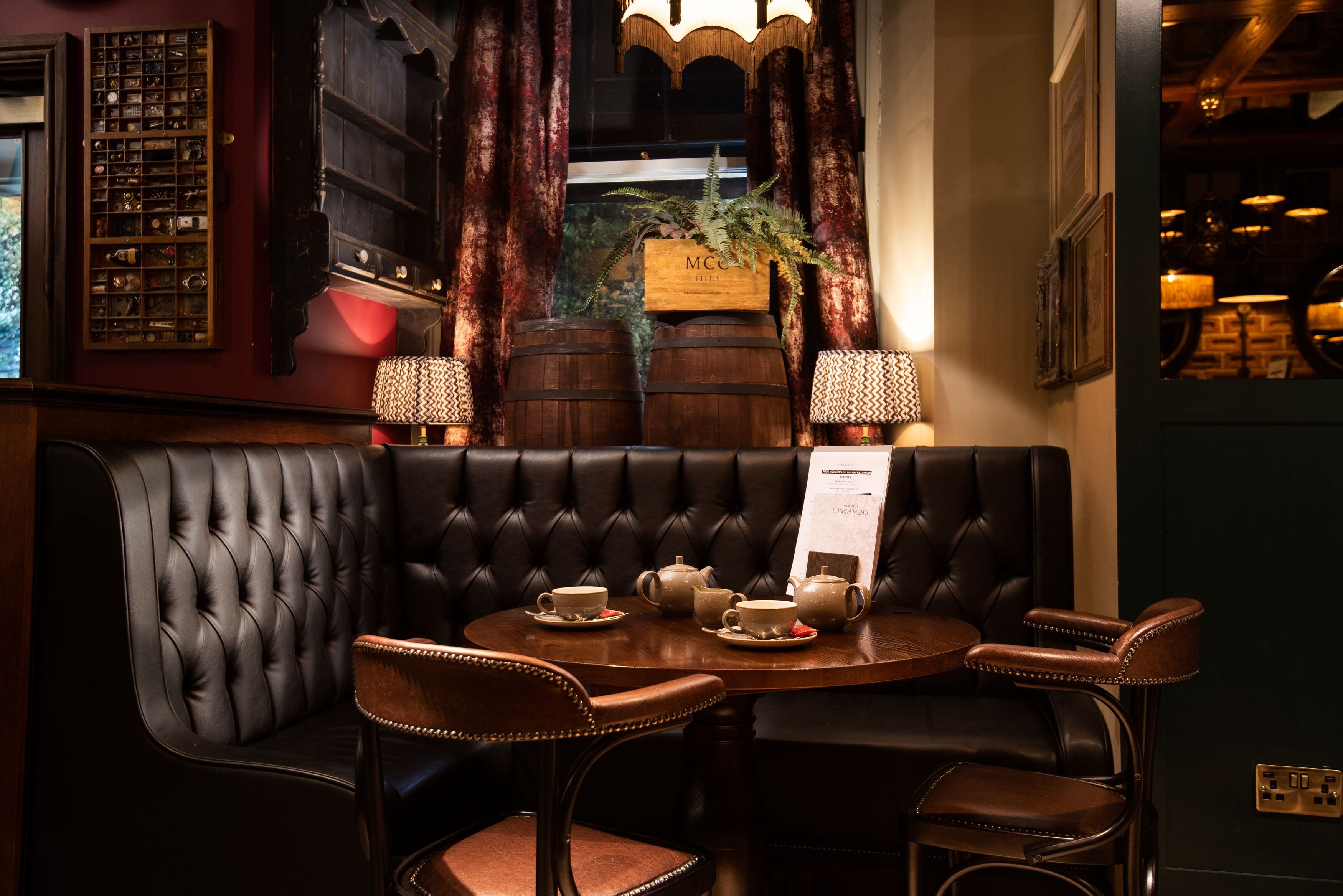 A dark leather booth with a table set for tea, surrounded by wooden chairs, in a warmly lit room with vintage decor.