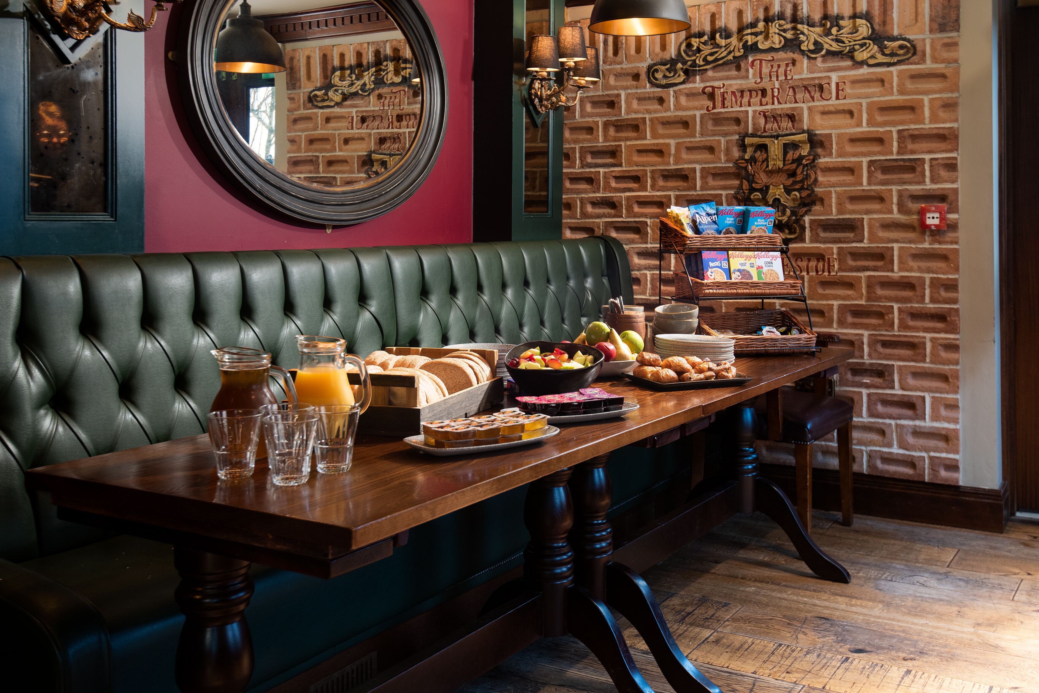 A dining area with a brick wall, a green upholstered bench, and a wooden table set with various breakfast items.