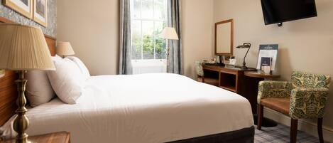 A hotel room with a large bed, a desk with a computer, a chair, a TV mounted on the wall, and a window with curtains.