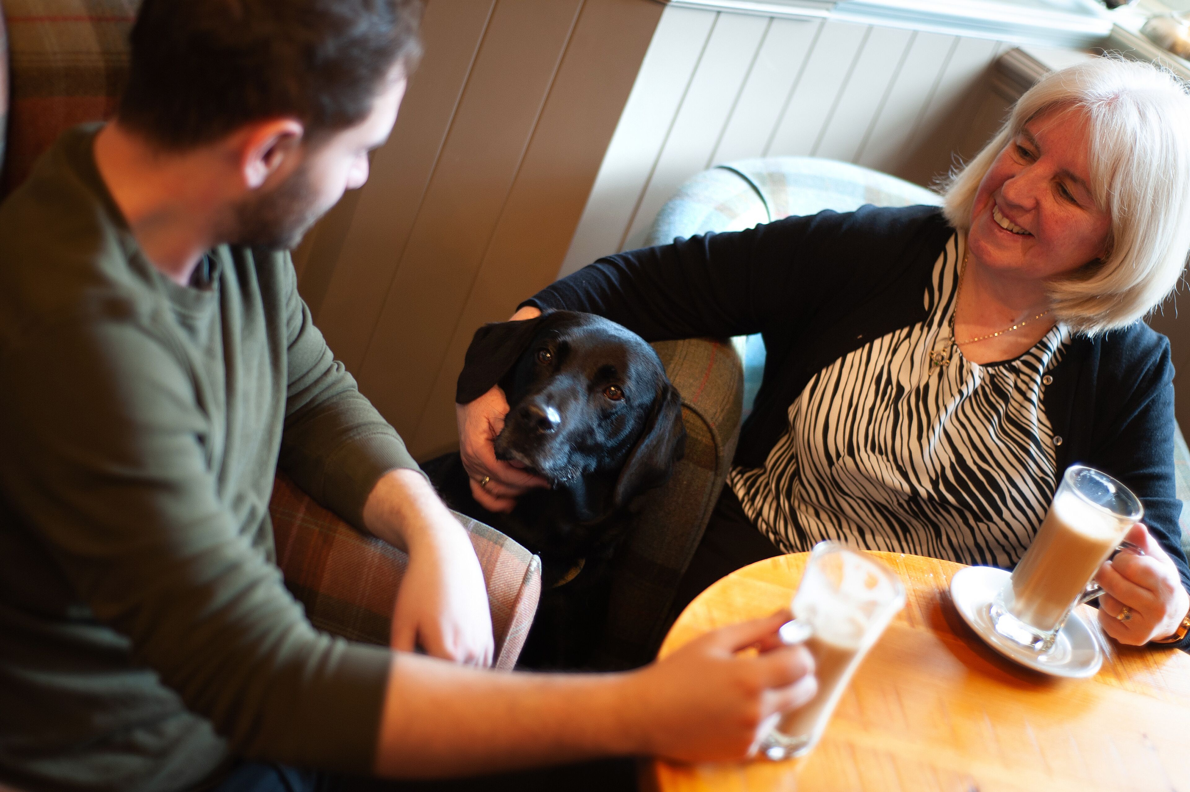 A person sitting at a table with a dog and holding a glass of a beverage.