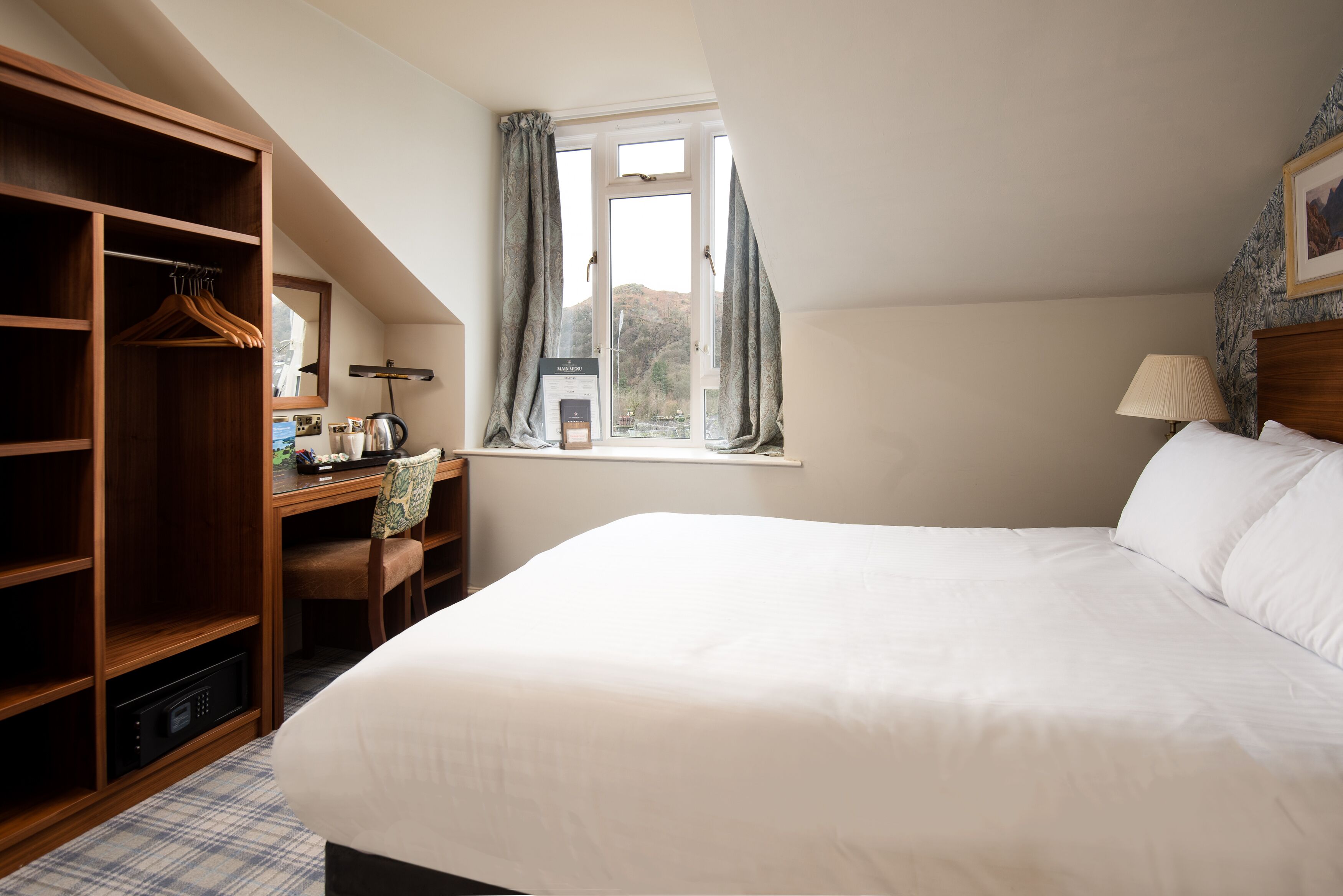 A bedroom with a large bed, a wooden wardrobe, a desk with a chair, and a window with curtains.
