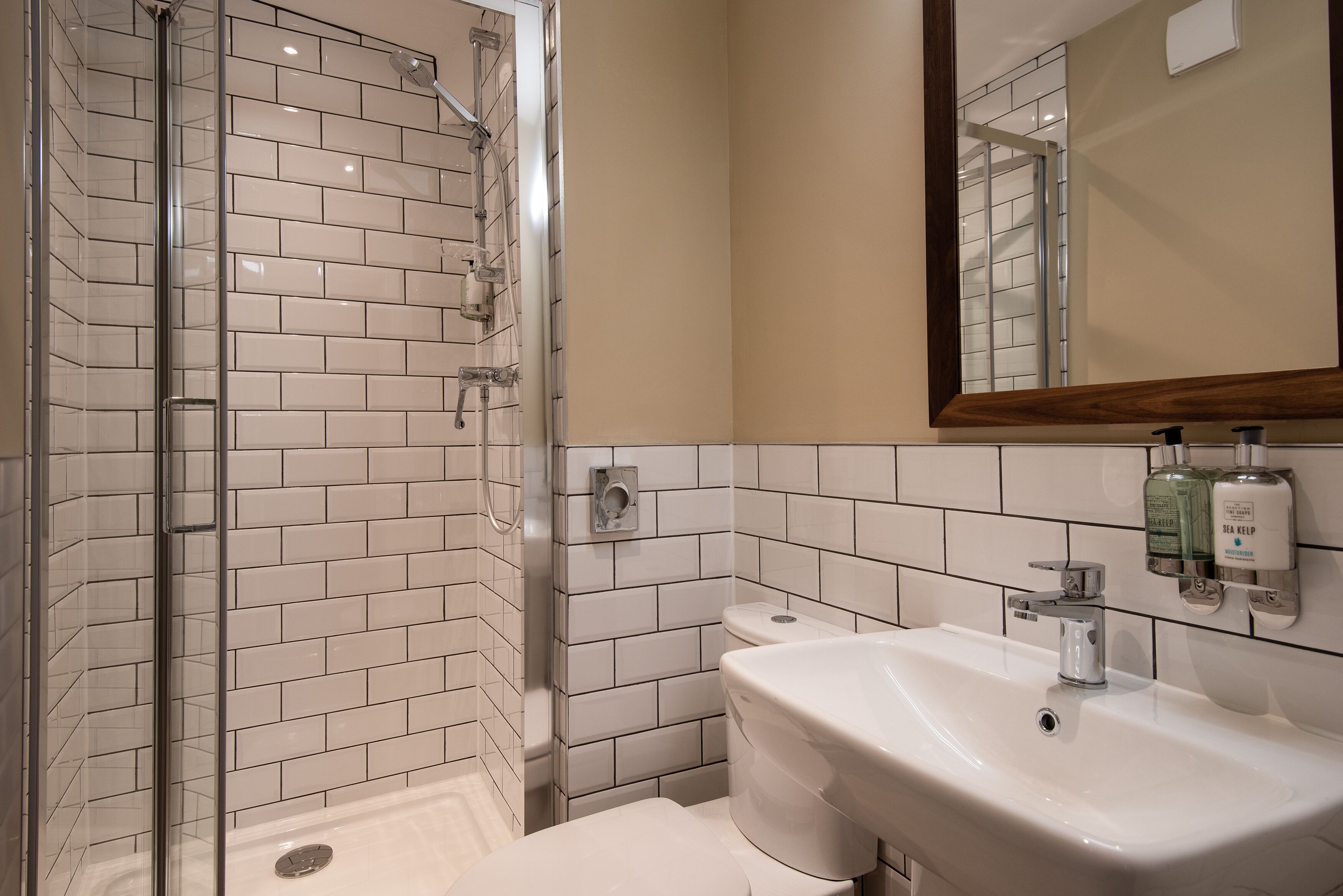 Une salle de bain avec une douche à parois de verre, des murs recouverts de carreaux blancs, un lavabo blanc et un miroir avec un cadre en bois.