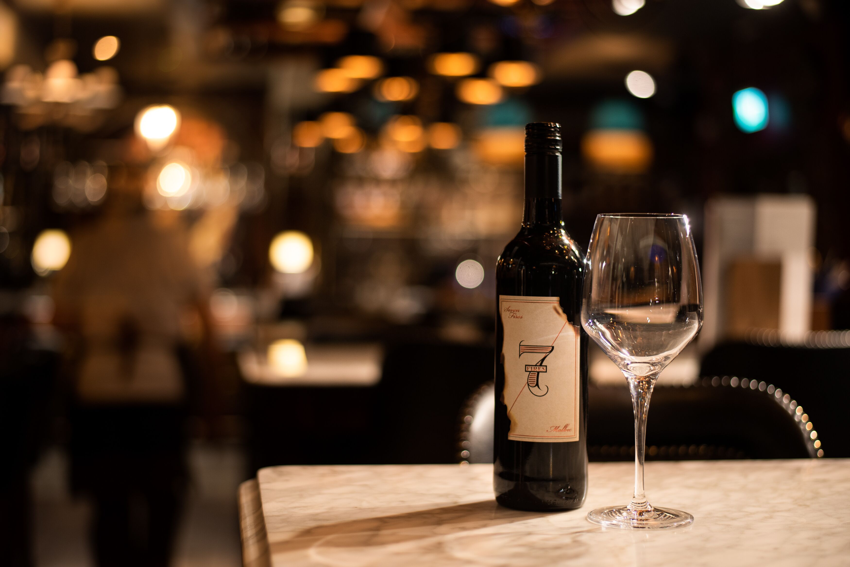 Una botella de vino y una copa vacía sobre una mesa en un bar con poca luz.