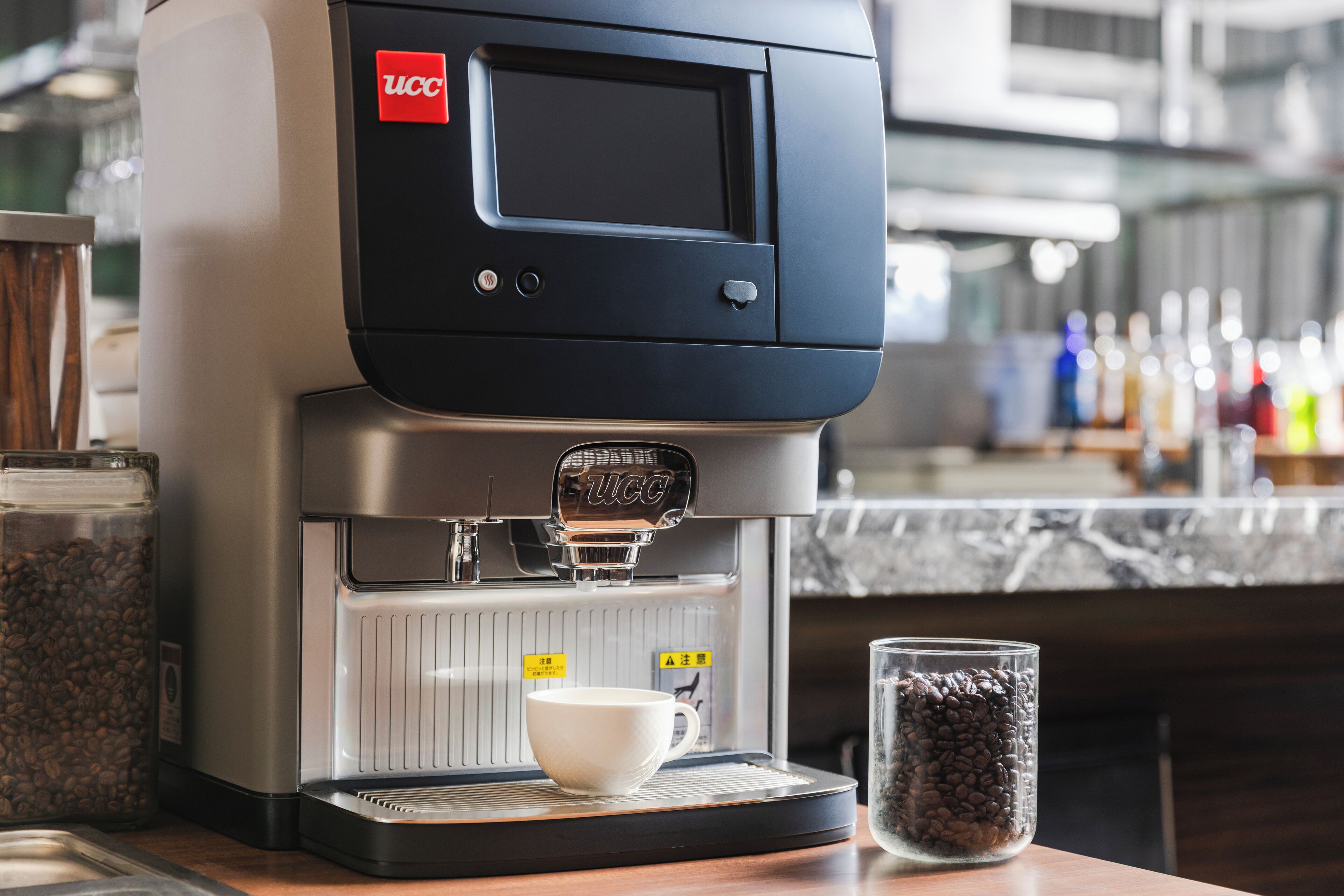 Una máquina de café comercial con pantalla digital, una taza de café y un recipiente con granos de café sobre una encimera.