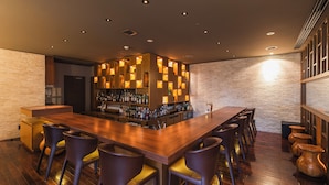 Eine Bar mit einer langen Holztheke, mehreren Barhocker und einer Wand mit beleuchteten Regalen, auf denen Flaschen präsentiert werden.
