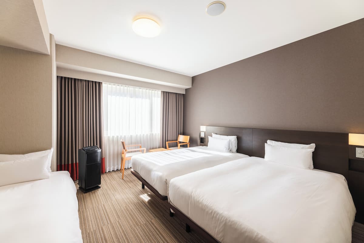 Une chambre d’hôtel avec deux lits, un bureau, une chaise, une poubelle et une grande fenêtre avec des rideaux.