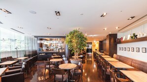 Ein modernes Restaurant mit Holztischen und -stühlen, einer zentralen Pflanze und einer Küche im Hintergrund.