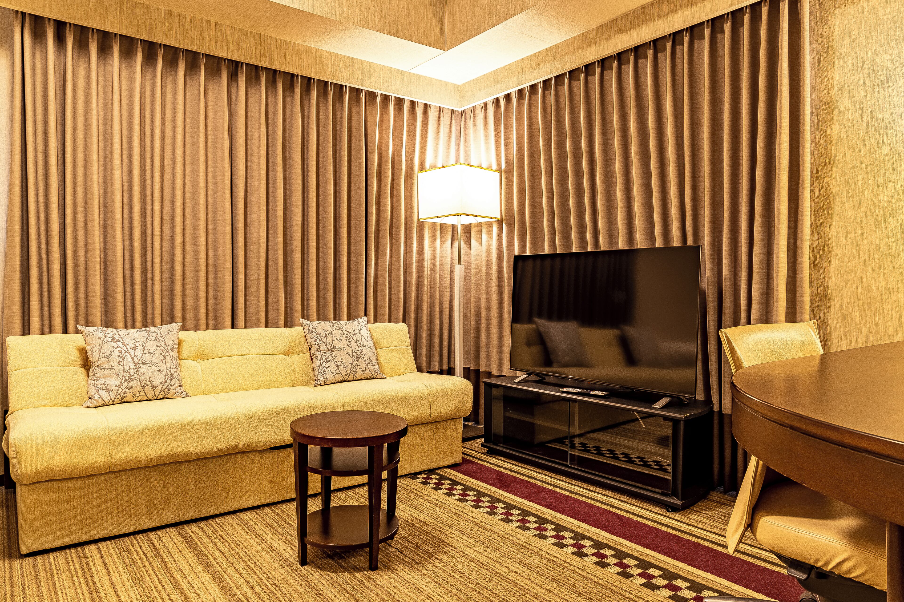 A hotel room with a yellow sofa, a TV, a small table, and a dining table.