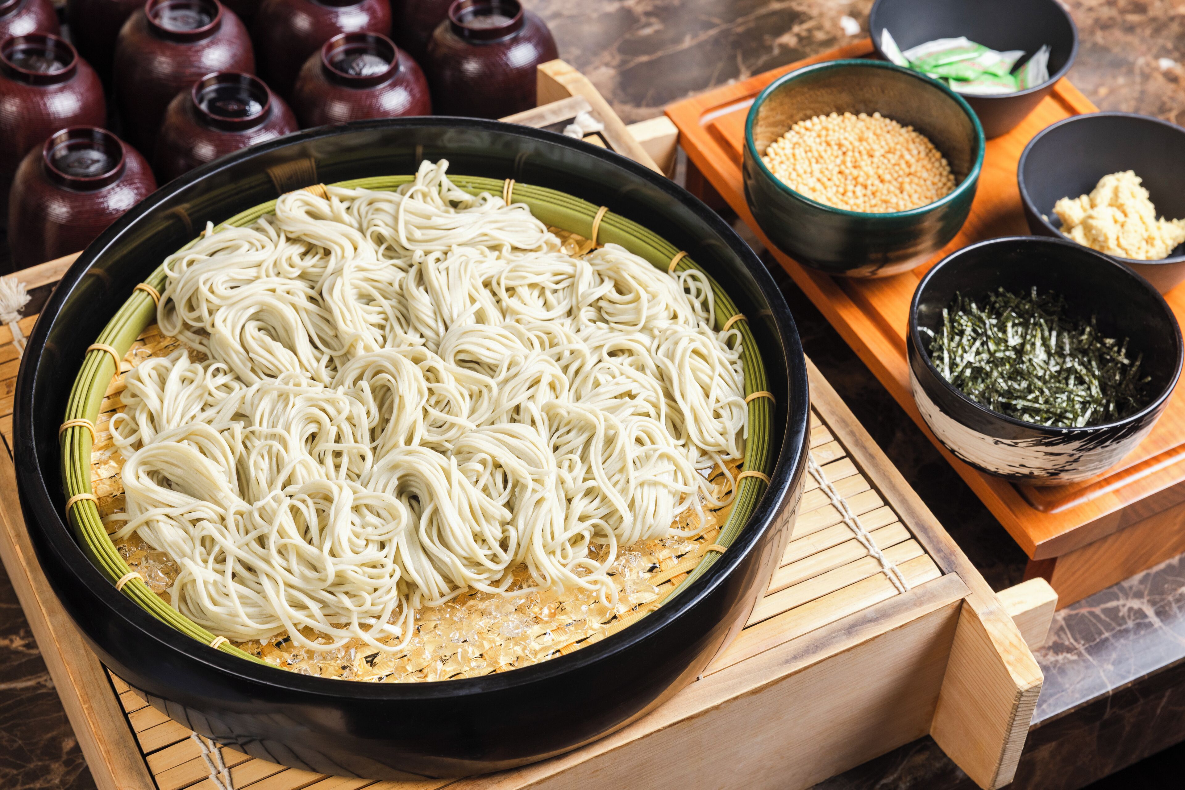 Un tazón de fideos blancos con acompañamientos variados, incluyendo verduras verdes y un tazón de semillas de sésamo.