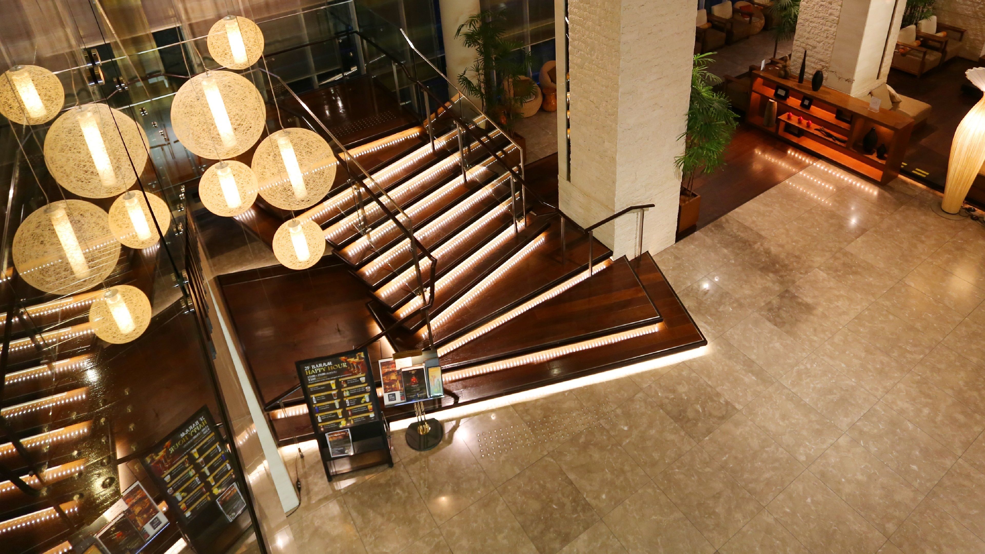 A modern hotel lobby with a wooden staircase, hanging spherical lights, and a reception desk.